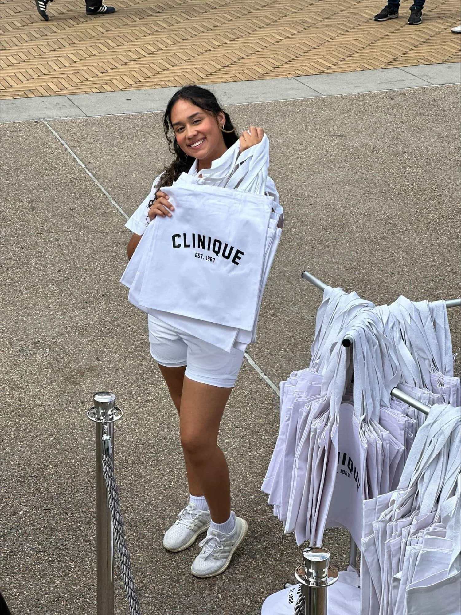 Young woman smiling, holding white tote bags with 'CLINIQUE' printed on them, standing outdoors next to a rack full of similar bags.