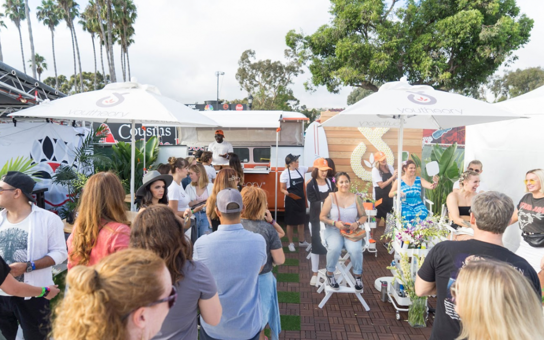 Crowd visiting an outdoor Youtheory brand activation popup featuring branded umbrellas, trailer bar, and beauty stations