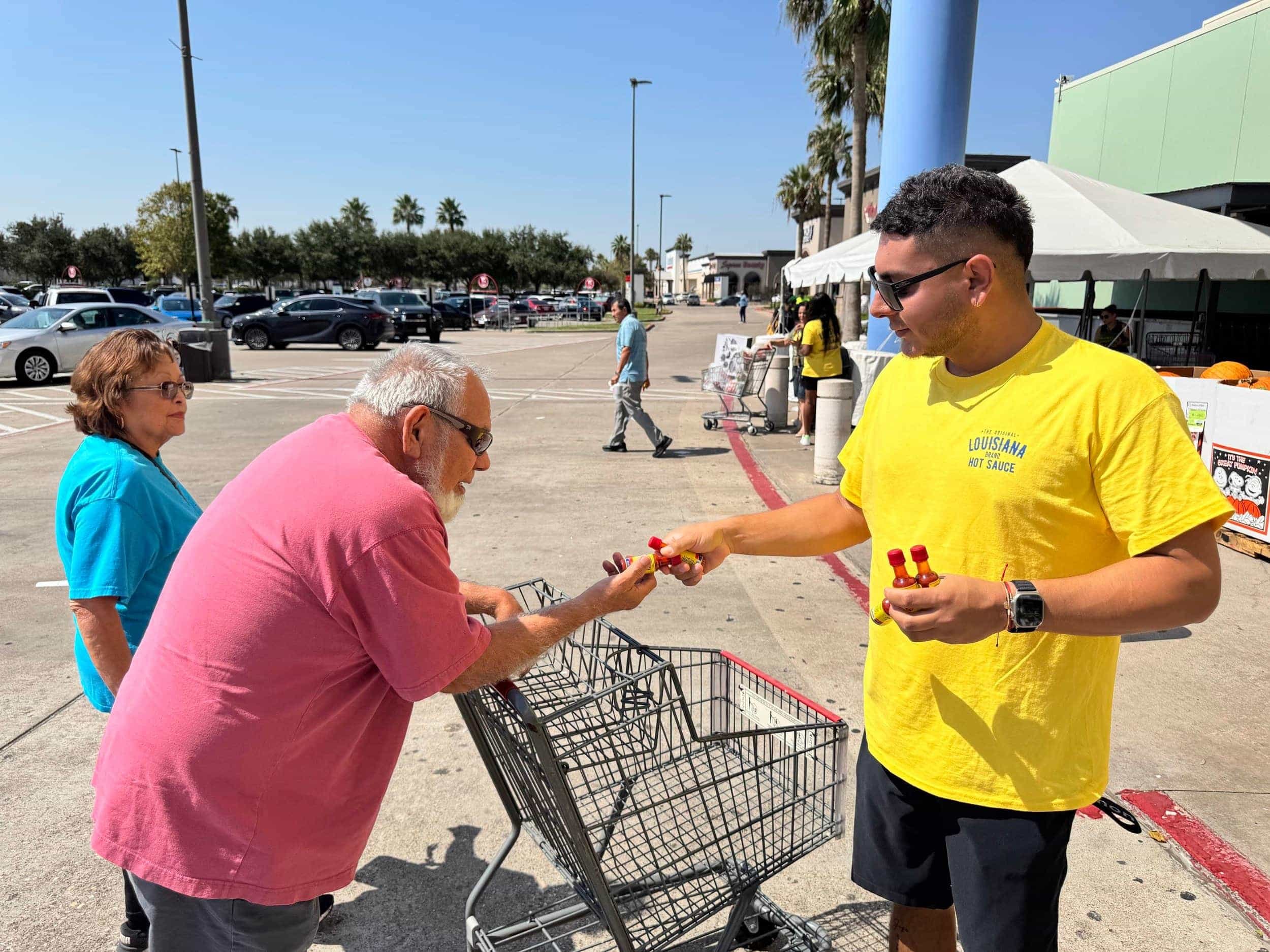 A man in a yellow T-shirt handing small hot sauce bottles to an elderly man in a pink shirt near a shopping cart in a parking lot while a woman in blue stands nearby under clear sunny skies.