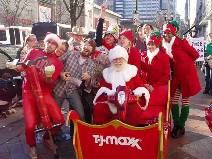 Santa Claus sitting in a TJ Maxx branded sleigh surrounded by festive event staff and promoters during an outdoor holiday parade - Encore Nationwide