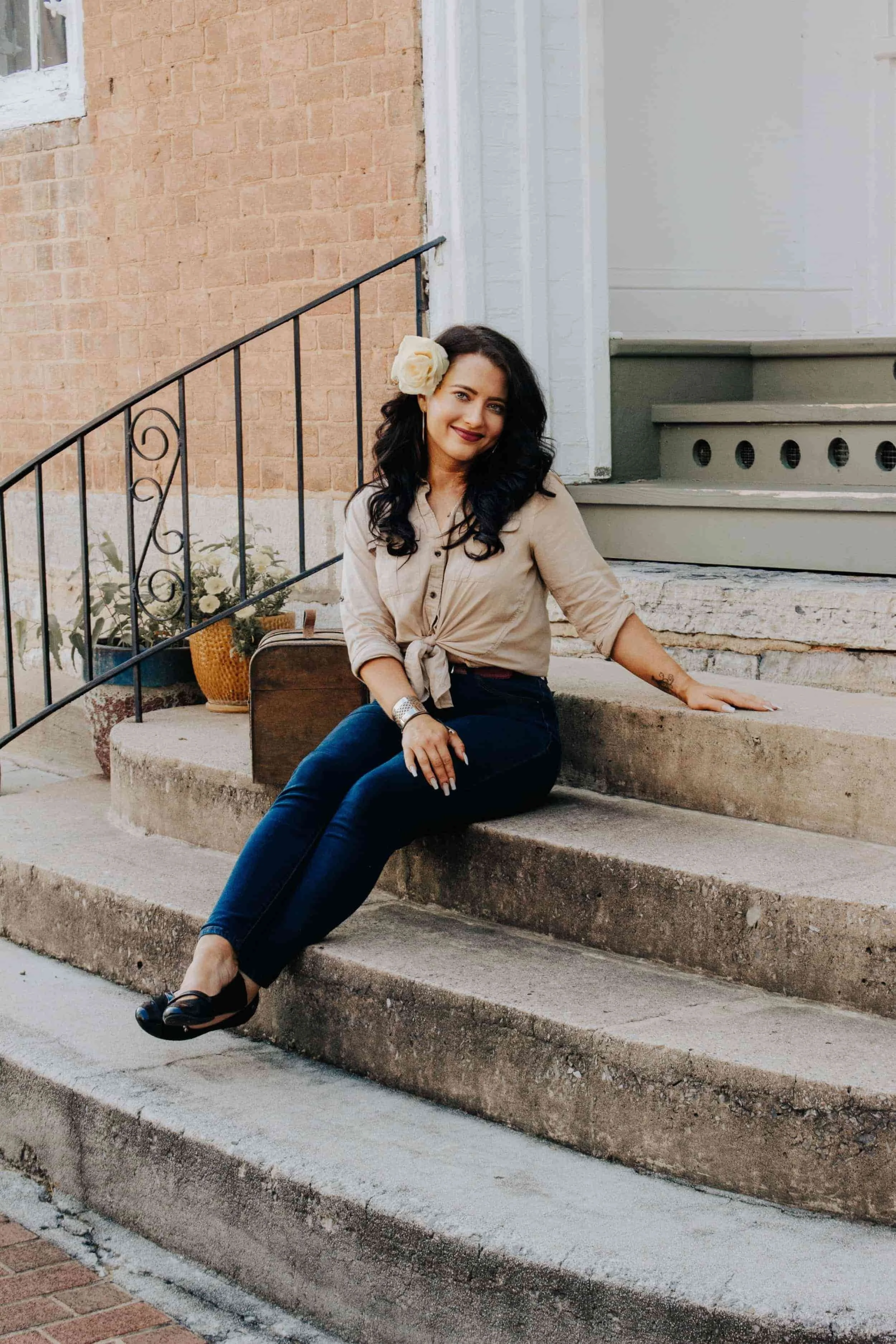 A woman sitting on outdoor stone steps, wearing a beige shirt tied at the waist, dark jeans, and black shoes, with a large cream-colored flower in her hair, smiling at the camera.