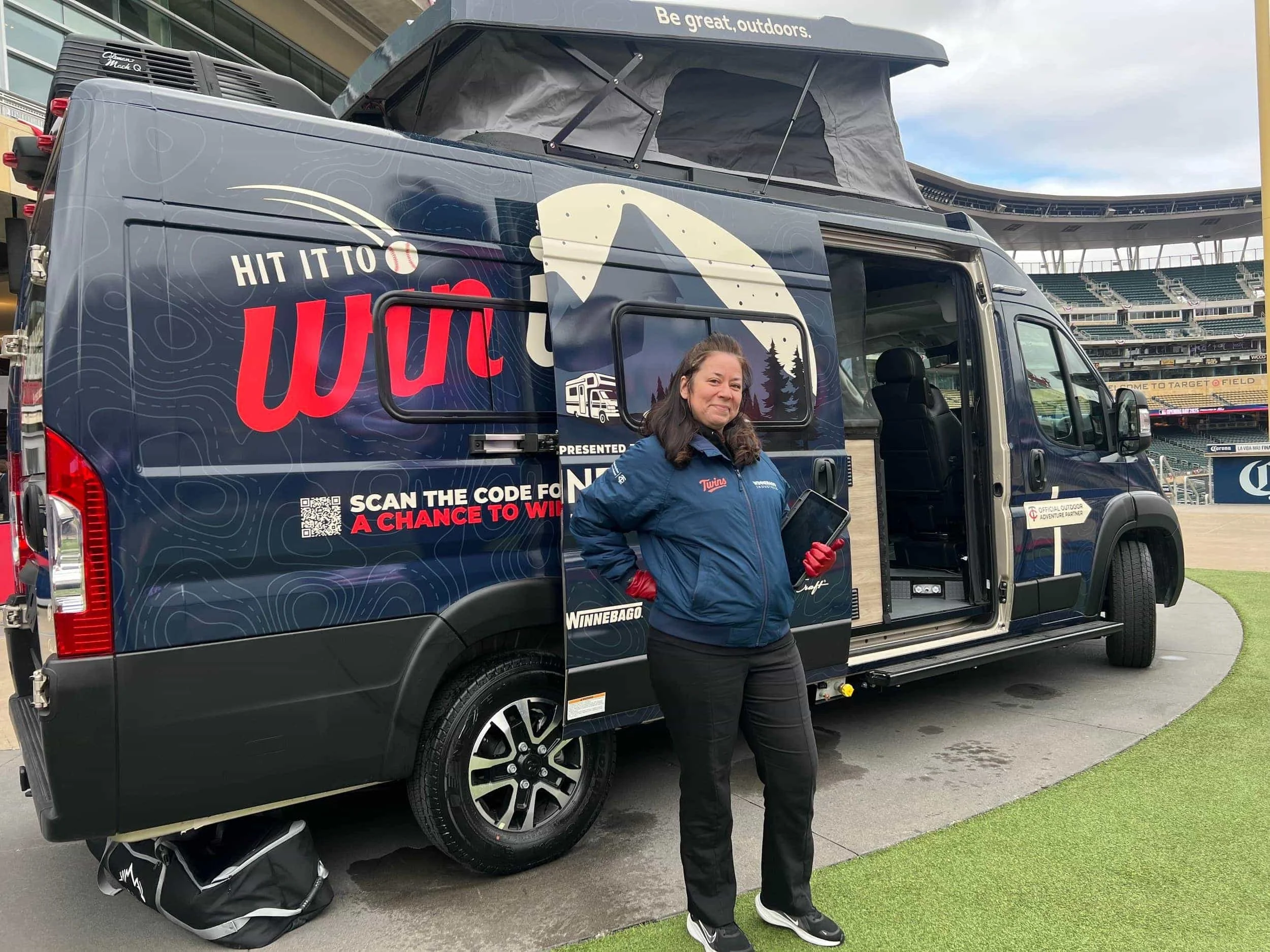 A woman standing outside a sports stadium next to a branded camper van, wearing a blue jacket, black pants, and sneakers, holding a tablet or clipboard, with a smiling expression.