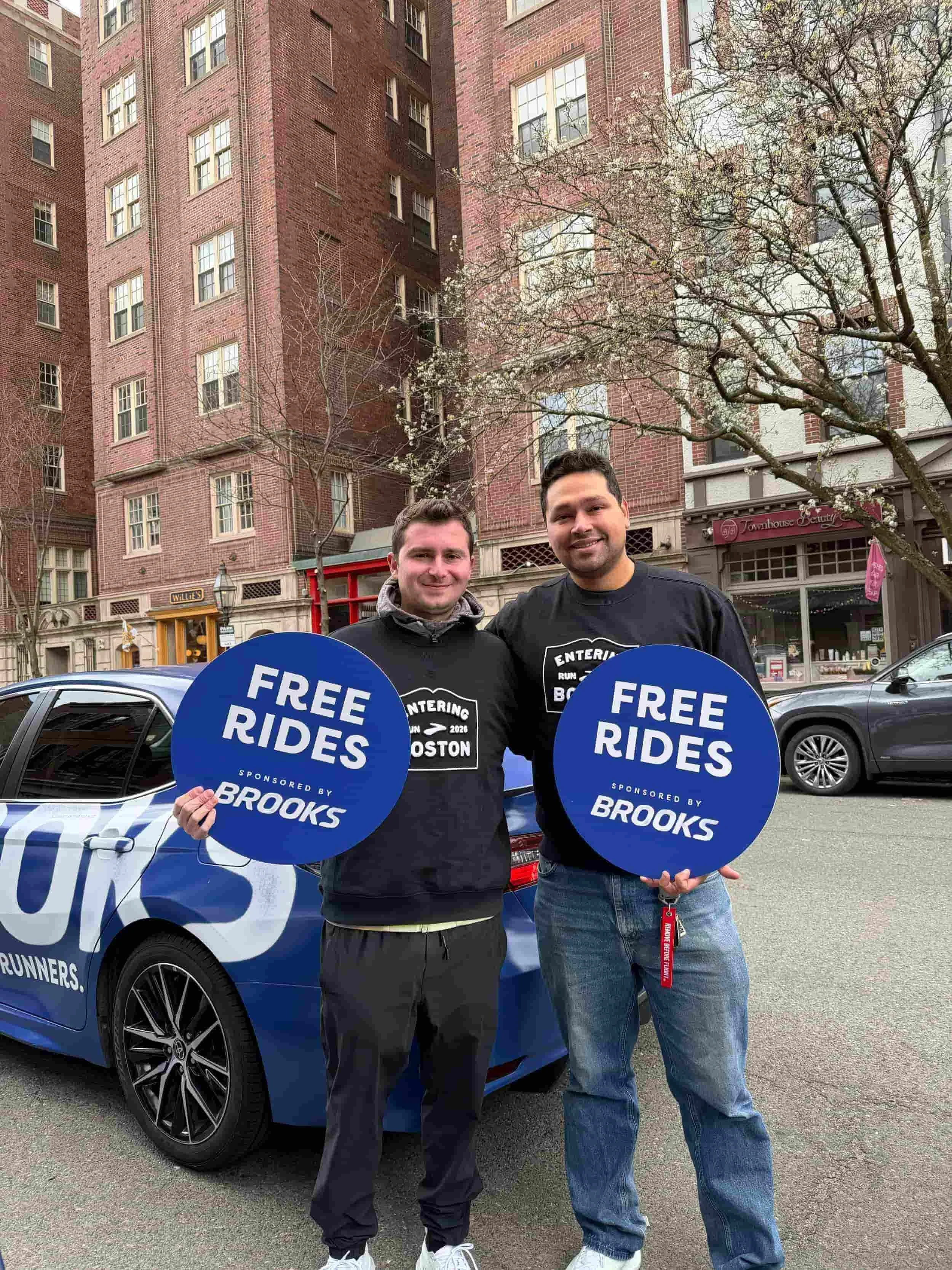 Two men holding blue 'Free Rides Sponsored by Brooks' circular signs standing in front of a branded Brooks car.