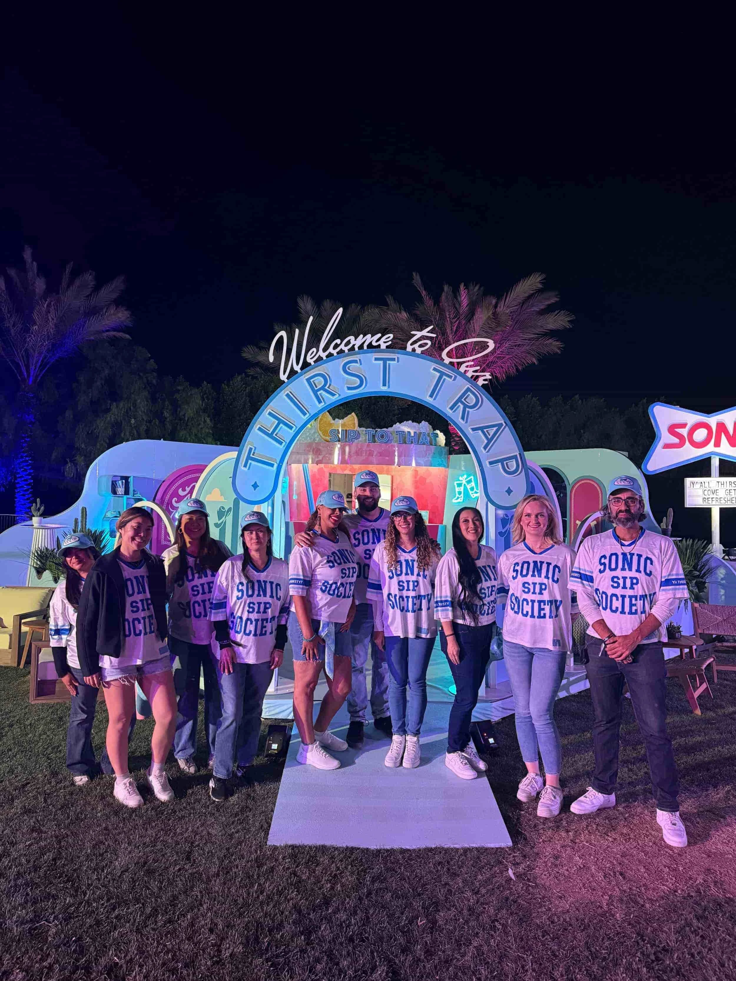 A group of staff wearing matching Sonic Sip Society jerseys posing in front of a illuminated Thirst Trap archway at night.