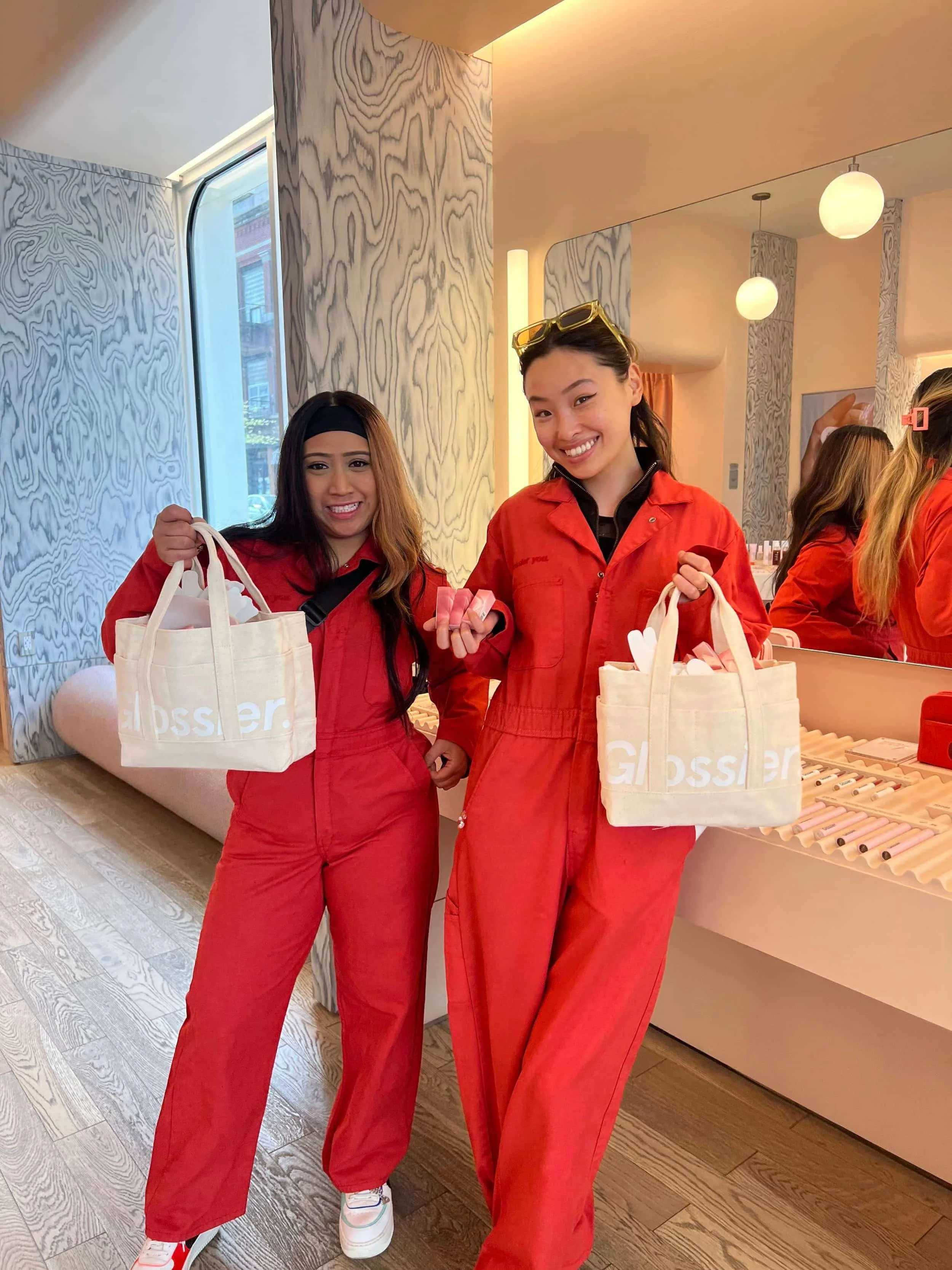 Two women wearing red jumpsuits are smiling and holding tote bags filled with items inside a modern, well-lit space with wood-patterned walls and a large mirror.