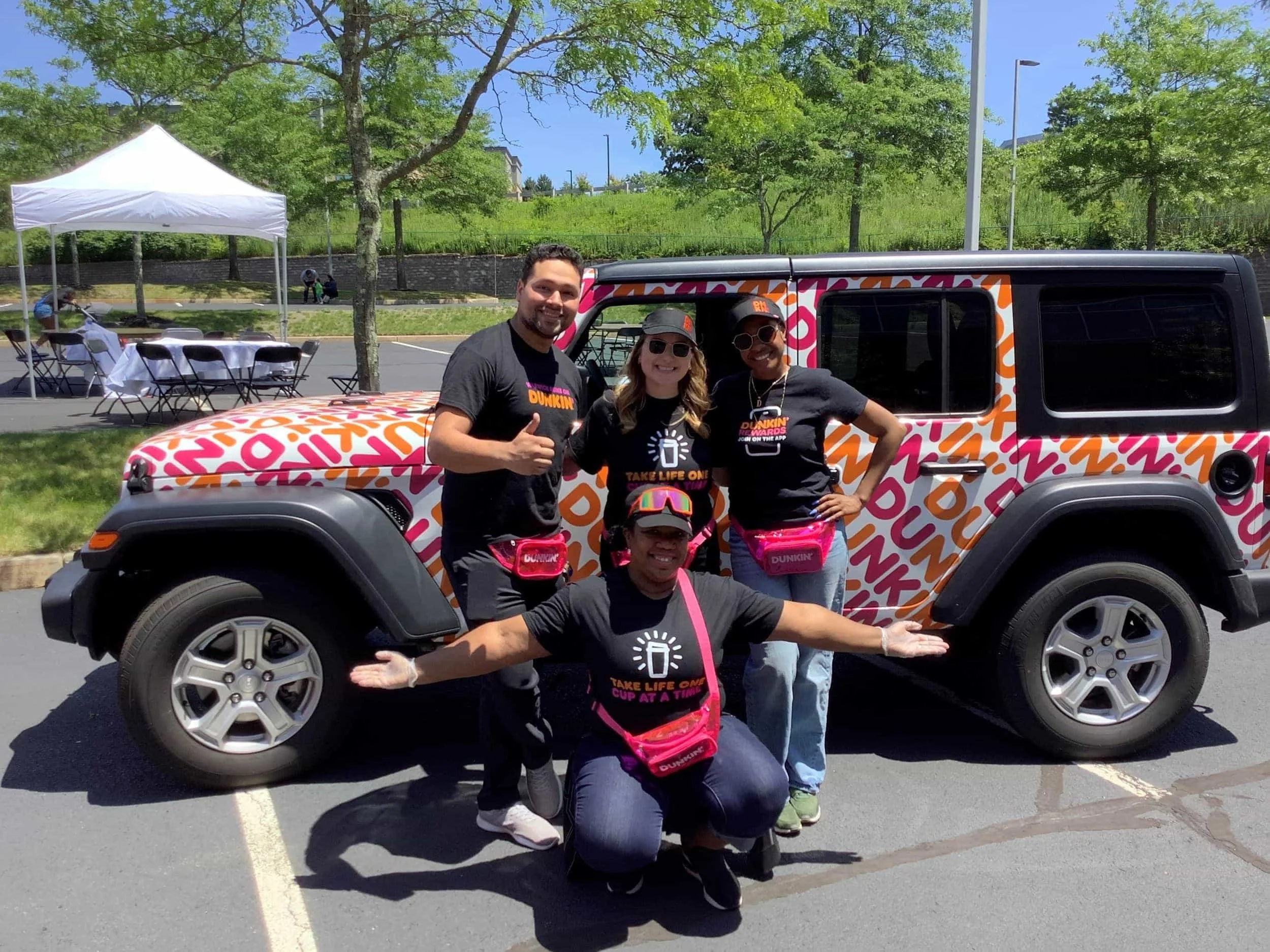 Four people posing in front of a decorated vehicle during a sunny outdoor event. They are wearing matching black shirts with colorful logos and pink fanny packs.