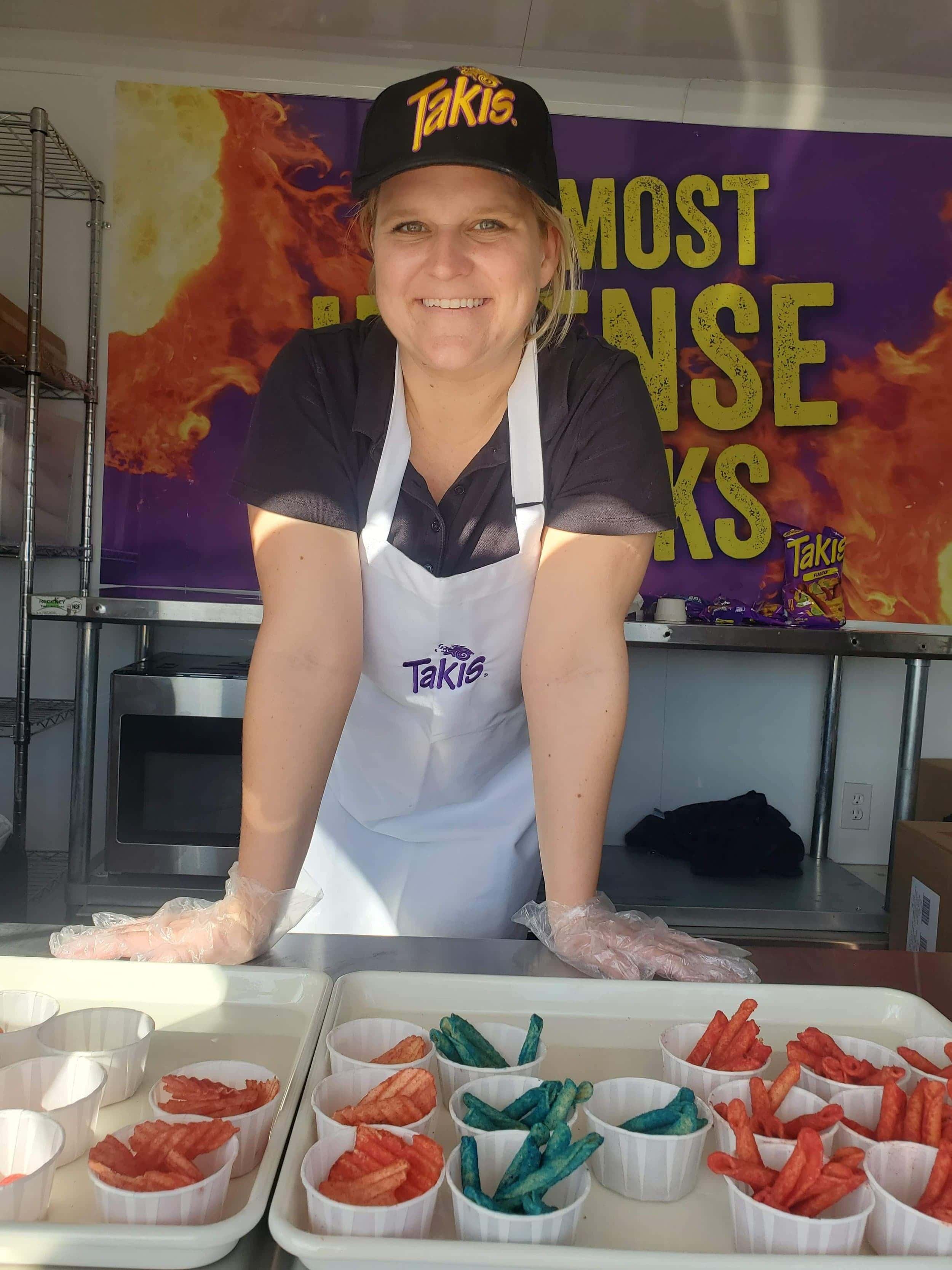 Smiling staff member wearing a Takis hat and apron standing behind sample cups of colorful Takis chips.