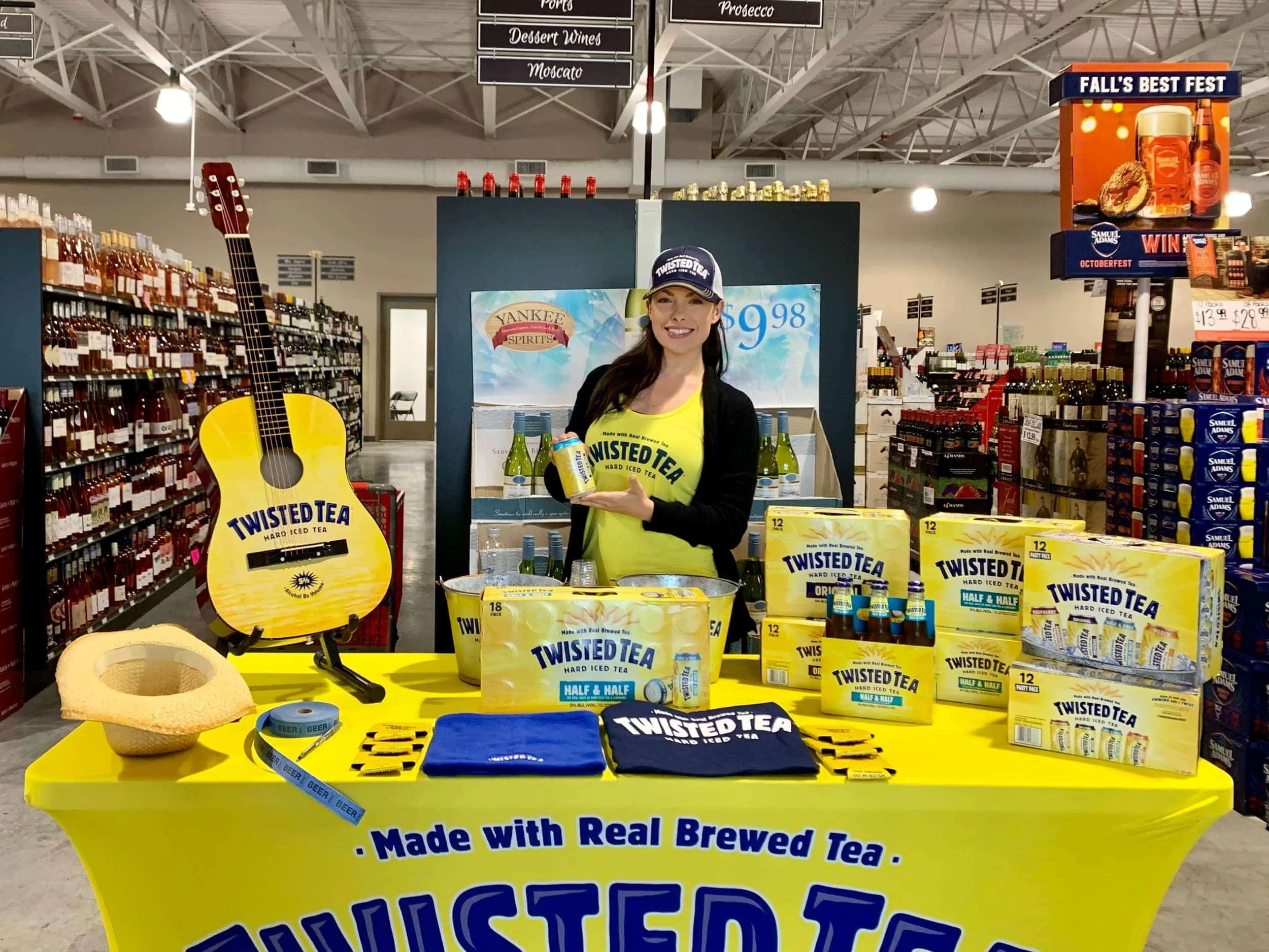 Brand ambassador standing behind a Twisted Tea promotional table display with branded merchandise and acoustic guitar in a liquor store