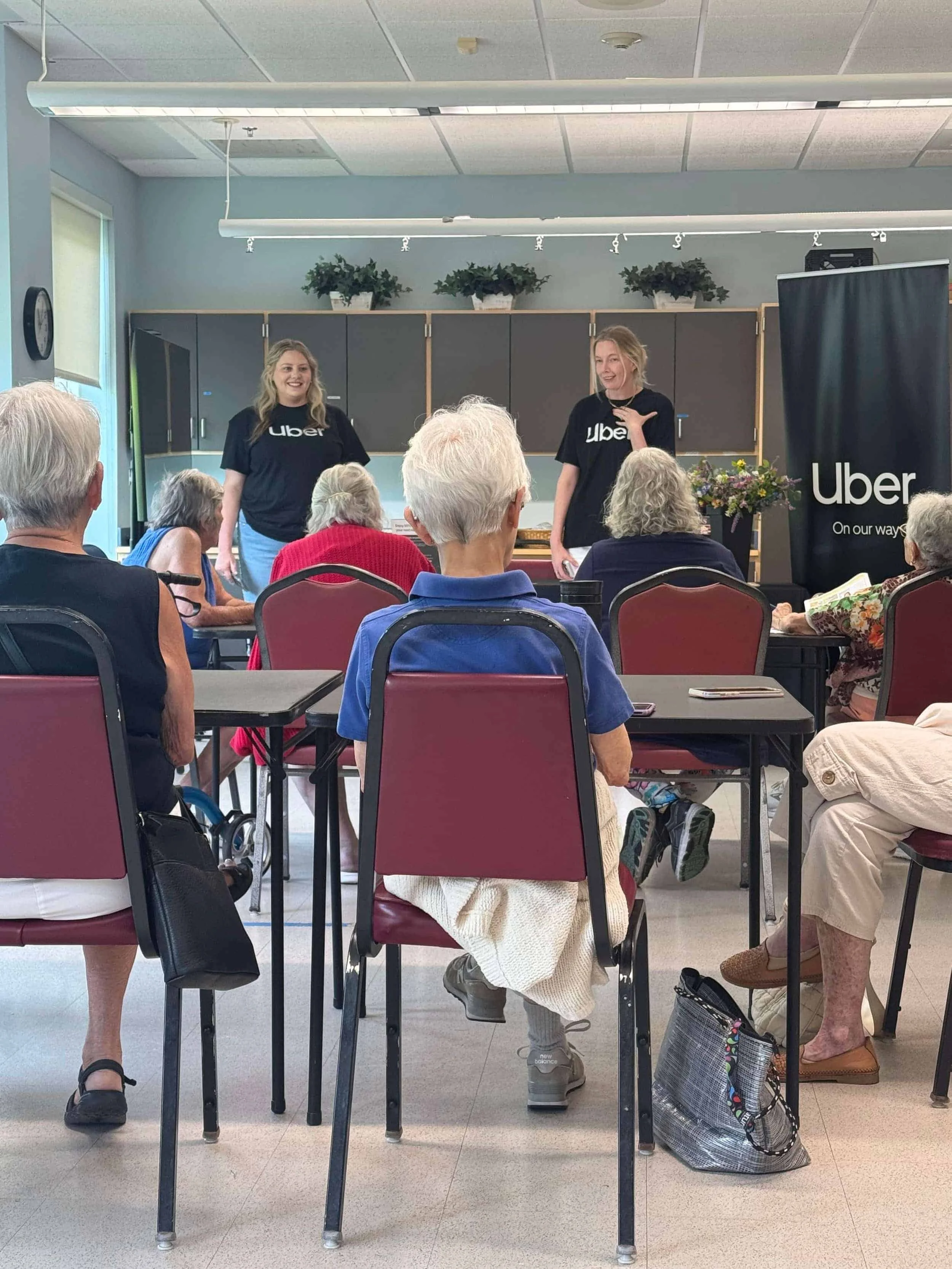 Two Uber representatives in black logo shirts giving an informational workshop presentation to an audience of senior citizens