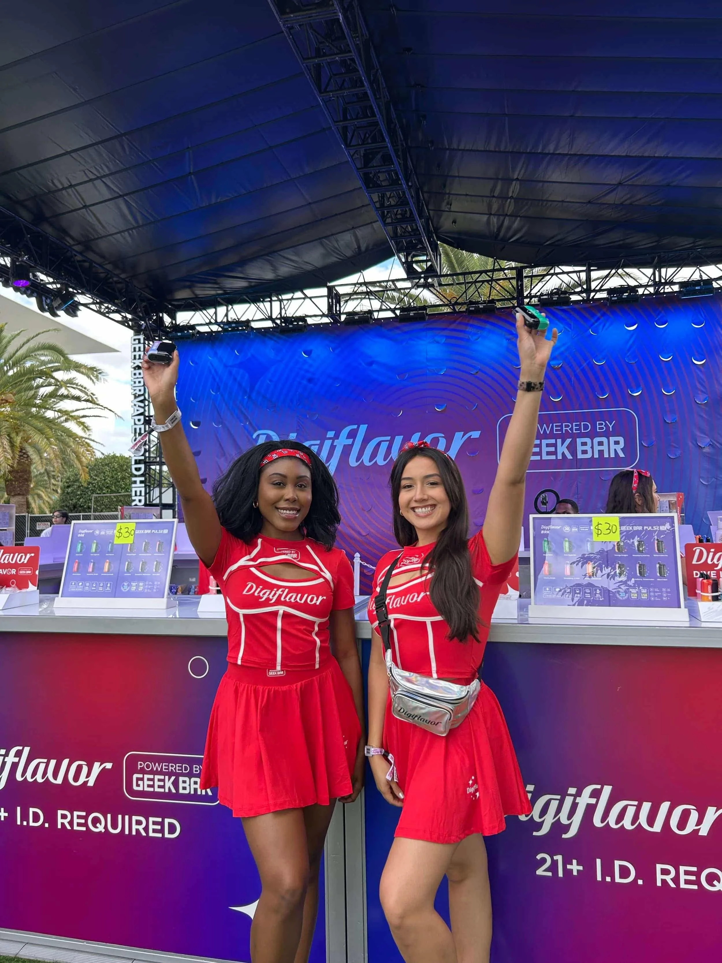 Two young women are standing in front of a DigiFlavor booth at an outdoor event, wearing red DigiFlavor branded outfits, smiling, and raising their hands. The booth has a blue and purple backdrop with promotional signage.