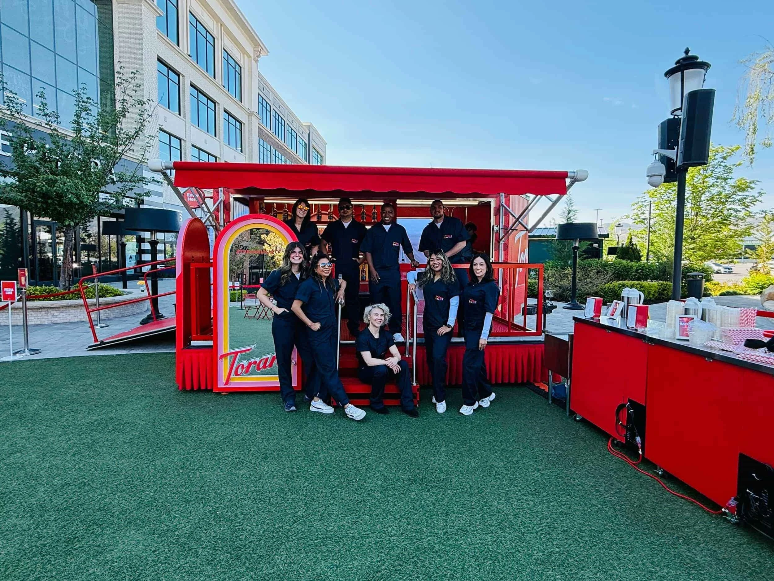 Event staff in matching dark uniforms posing around a red Torani mobile bar trailer at an outdoor pop-up event