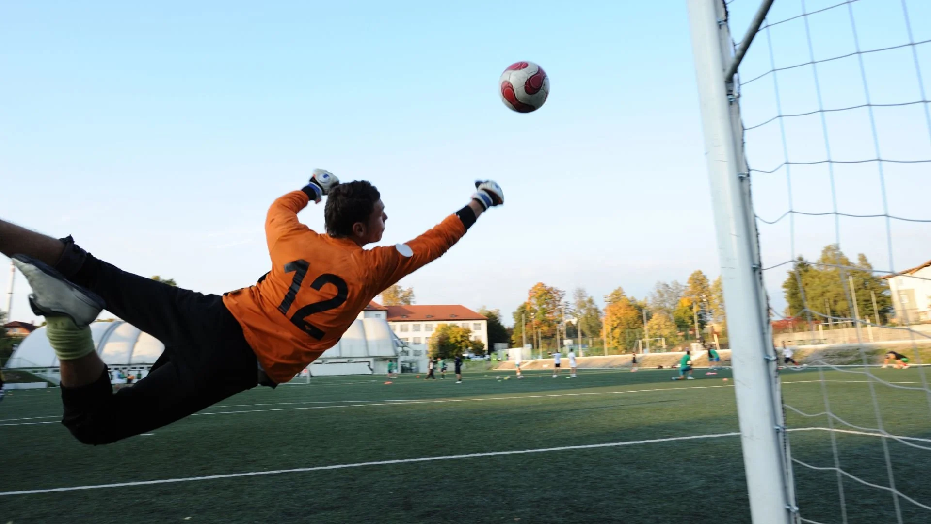 A soccer goalkeeper in an orange jersey diving in mid-air to save a ball near the goal post