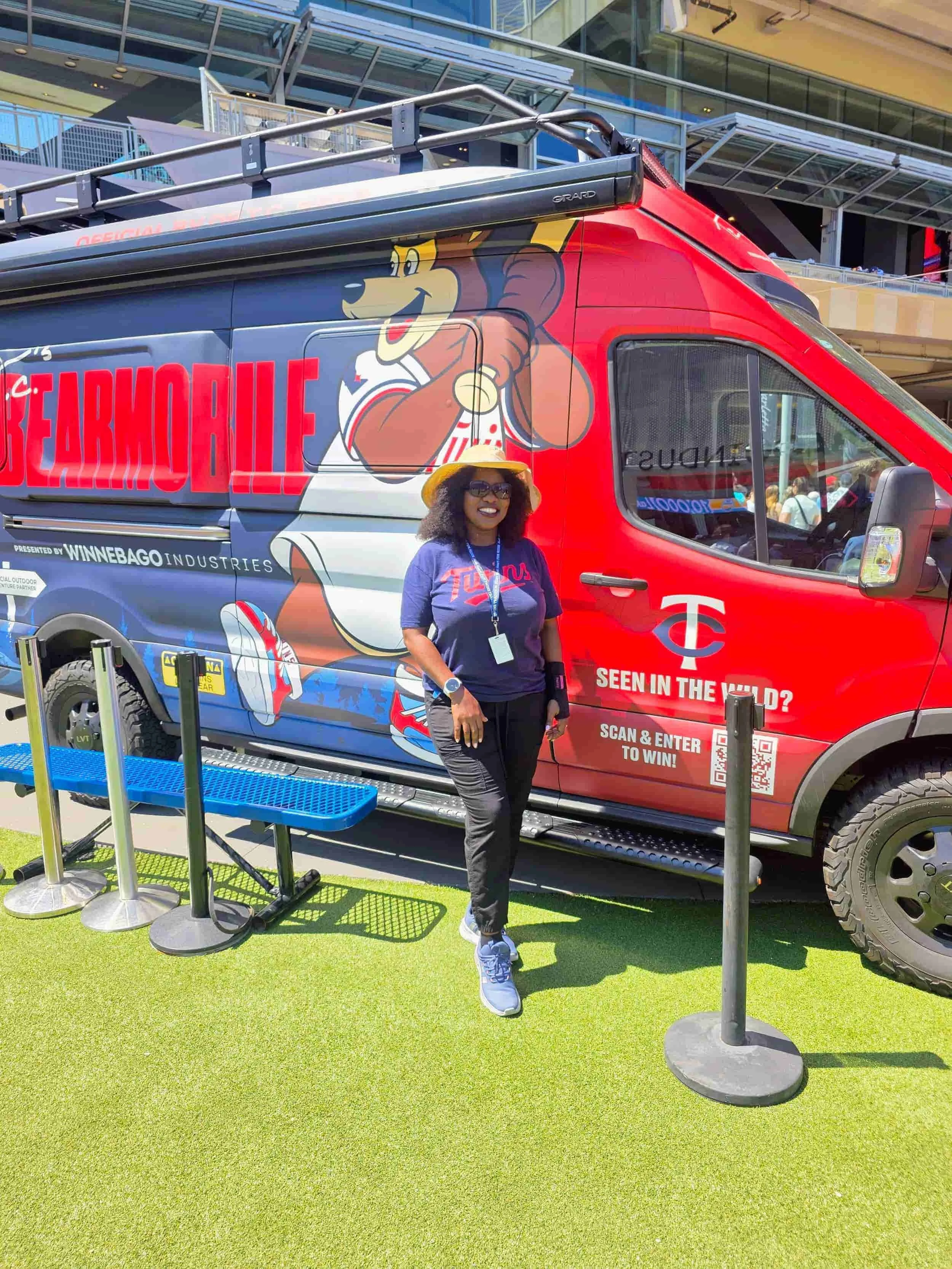 A woman wearing a Minnesota Twins shirt and straw hat posing in front of a Minnesota Twins Bearmobile camper van