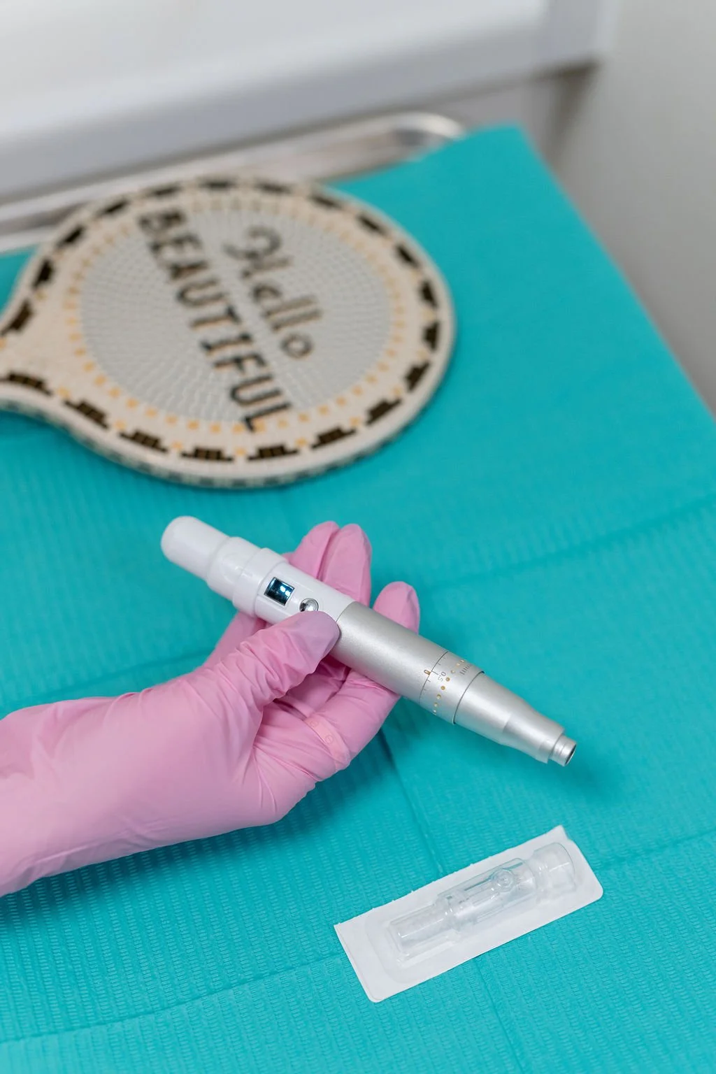 A hand wearing a pink glove holding a handheld medical device, possibly a laser or plasma pen, on a teal medical surface. In the background, there is a woven circular sign that says "The Future" and part of a blister pack with a small glass vial.