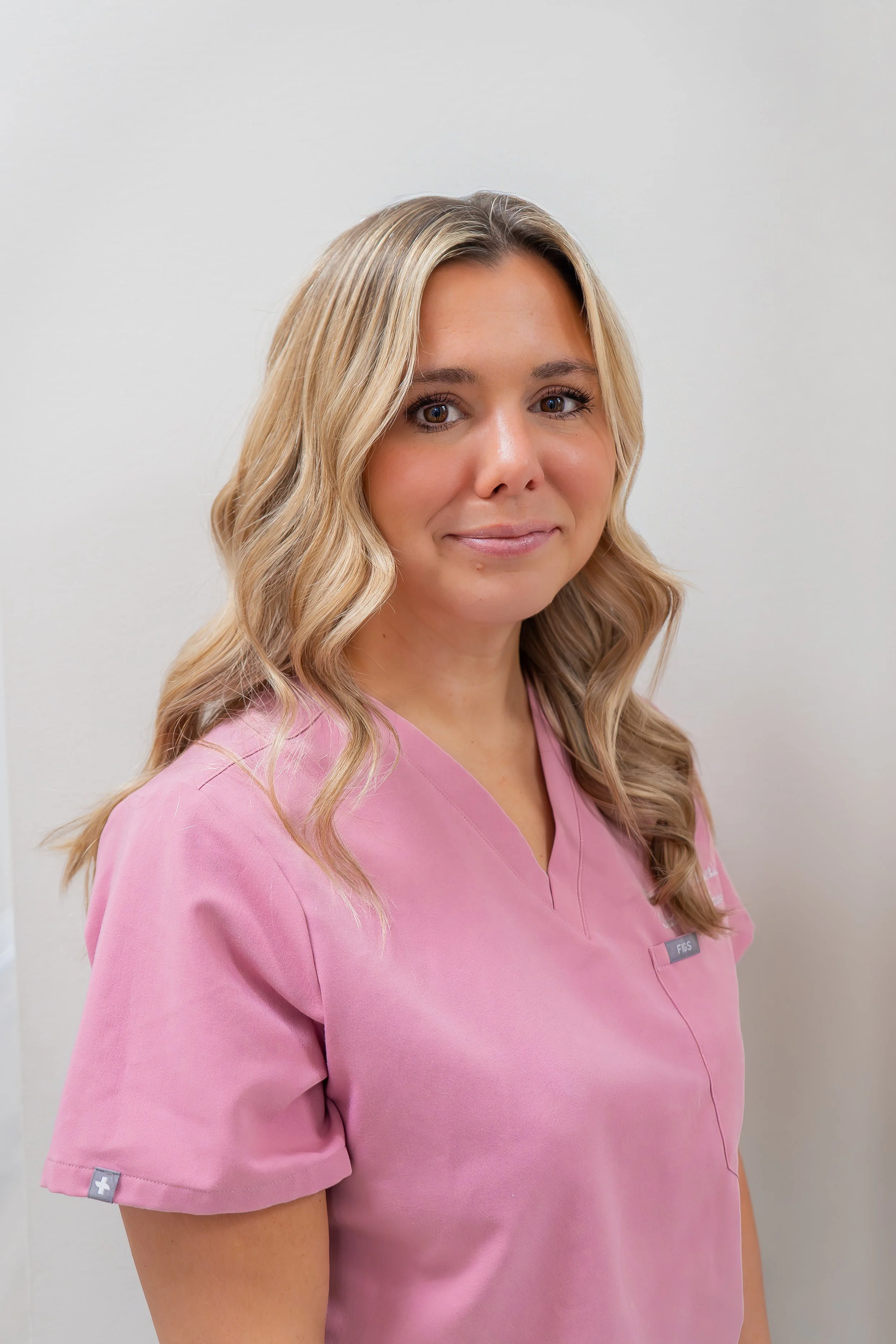 A woman with blonde hair styled in loose waves, wearing pink medical scrubs and standing against a neutral background.