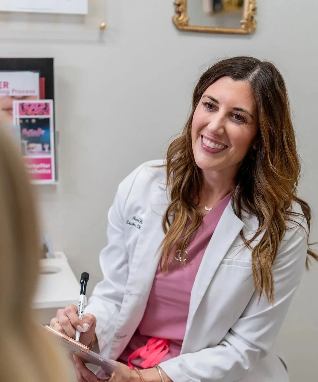 A female CRNA with long brown hair, wearing a white coat and pink scrubs, smiling and holding a clipboard, in a medical office setting.