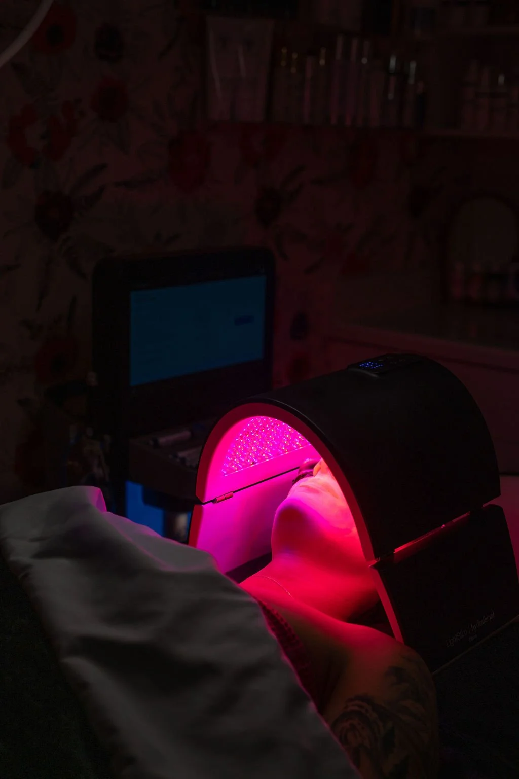 A person is undergoing a red light therapy treatment using a specialized device that emits pinkish-red light onto their face.