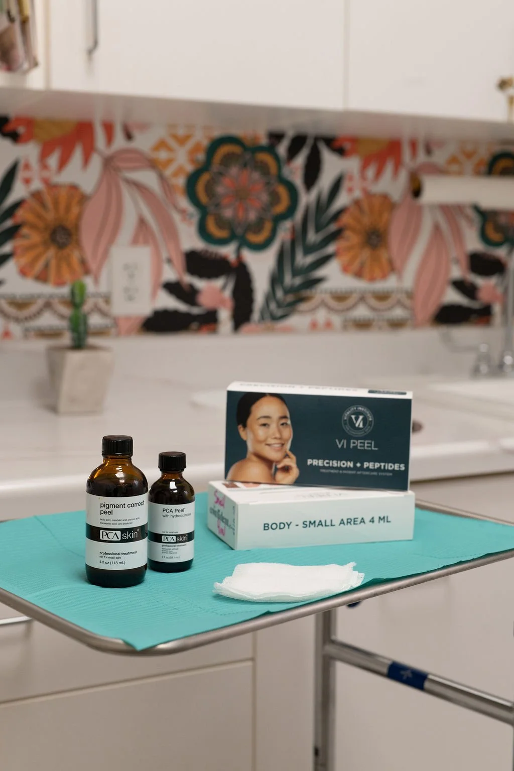 Medical treatment setup with skincare products including PCA Skinpigment Correct Peel and PCA Peel, on a teal drape with a white tissue, in a clinical room with colorful floral patterned wallpaper in the background.