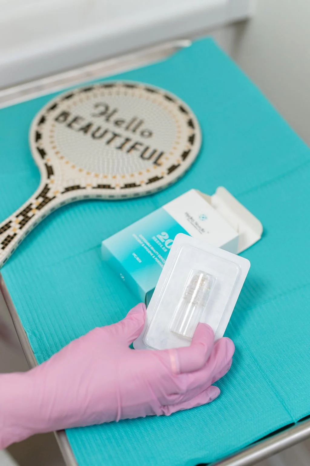 A person's hand wearing a pink glove holding a small sterile vial over a blue medical tray. There is a box of exosomes and PDGF kits and a sign shaped like a paddle that says 'Hello Beautiful' on the tray.