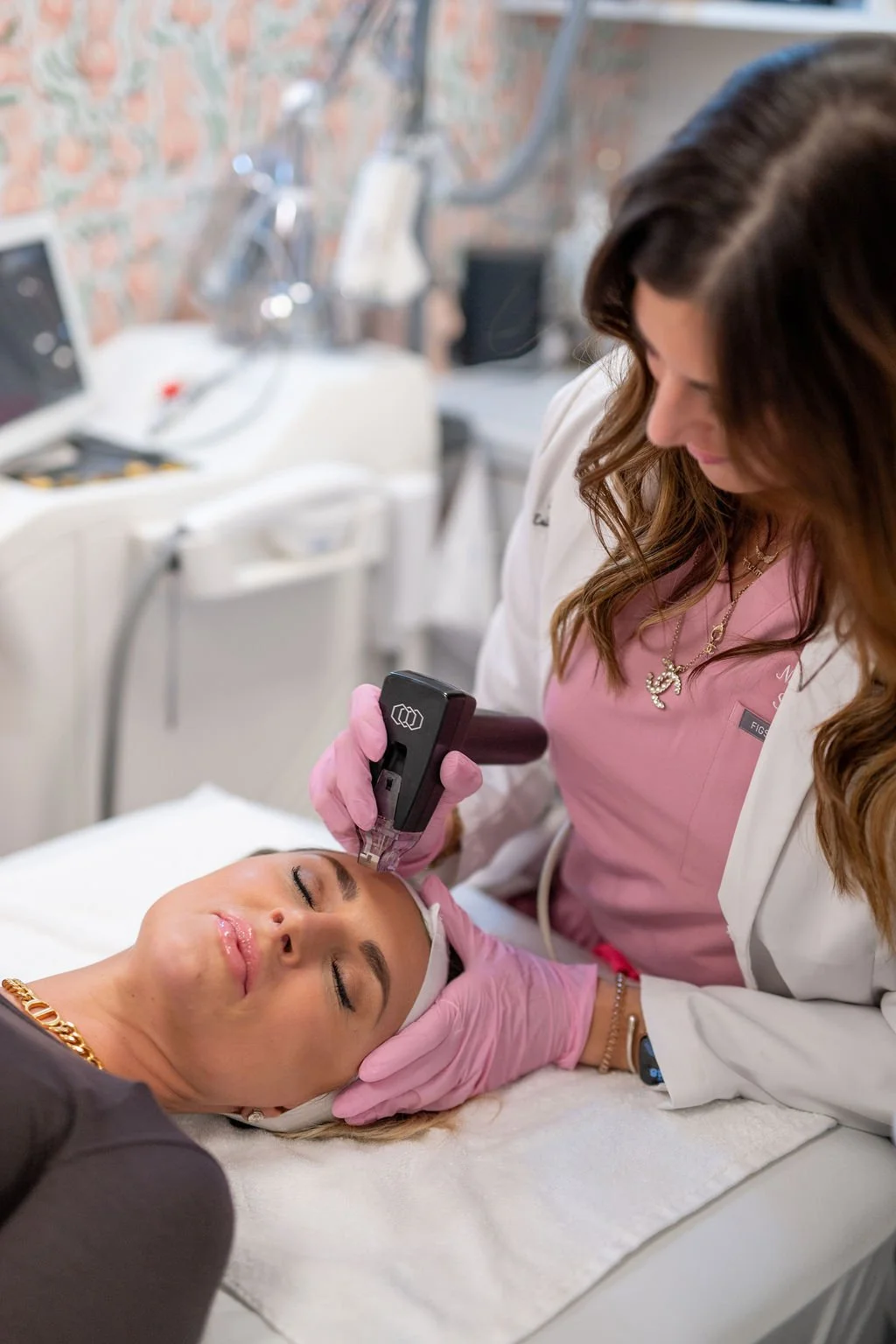 A woman receiving a Morpheus8 treatment with a device on her face in a beauty clinic. The CRNA practitioner is wearing pink gloves and a white coat, while the patient has a white headband and appears relaxed.