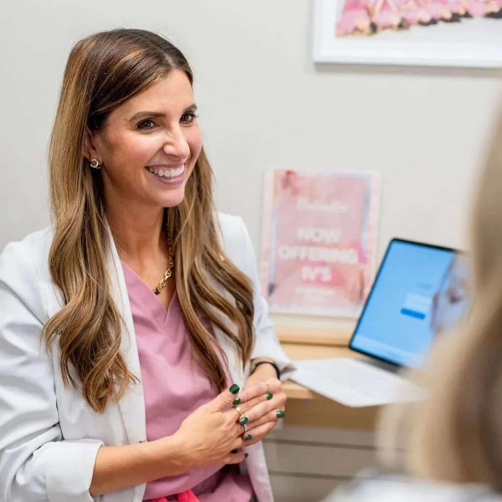 A nurse practitioner with light brown hair and a white coat talking to someone in an office setting.