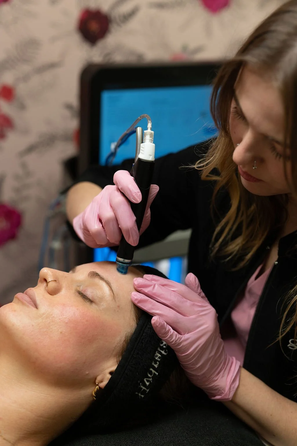 A woman receiving a Hydrafacial treatment with a device from a esthetician wearing pink gloves in a spa or clinic setting.