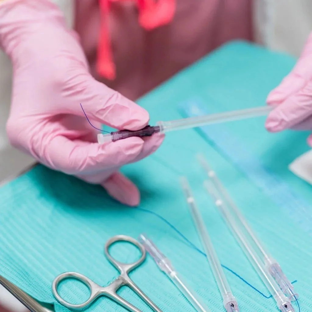 Close-up of threads with pink-gloved hands handling a syringe and needle on a teal surgical drape, with scissors and other medical tools nearby.