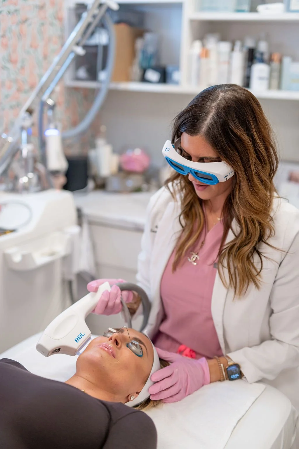 A woman lying on a treatment bed wearing eye protection glasses receives a Moxi facial treatment with a handheld device from a skincare professional wearing glasses, pink gloves, and a white coat in a clinic setting.