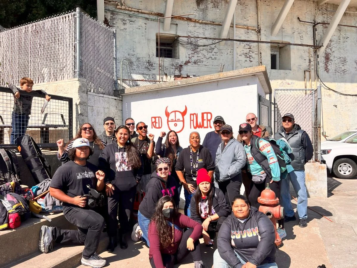 A group of people posing outdoors in front of a white wall with graffiti of a red devil's face and the words 'Red Rebel'. They are smiling, some with fists raised, and some are wearing sunglasses and casual clothing. There are backpacks and a camera 