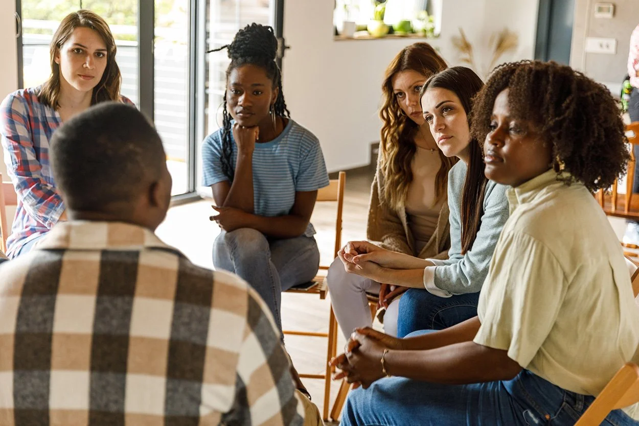 A diverse group of six women sitting in a circle having a discussion in a well-lit room with large windows.