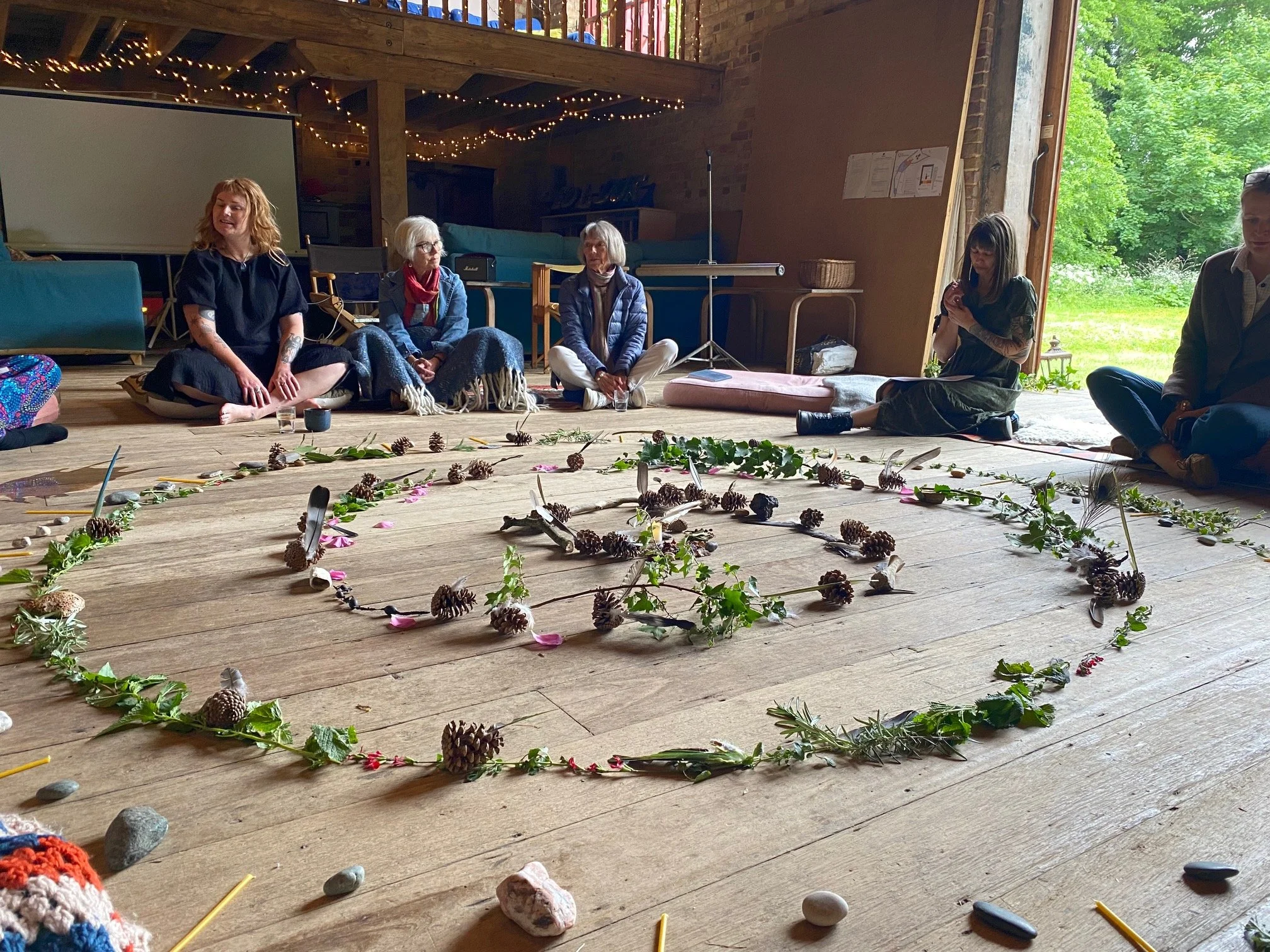 A group of people sitting on the floor in a circle inside a rustic room with wooden floors, surrounded by natural items like pinecones, leaves, and stones arranged in a pattern.