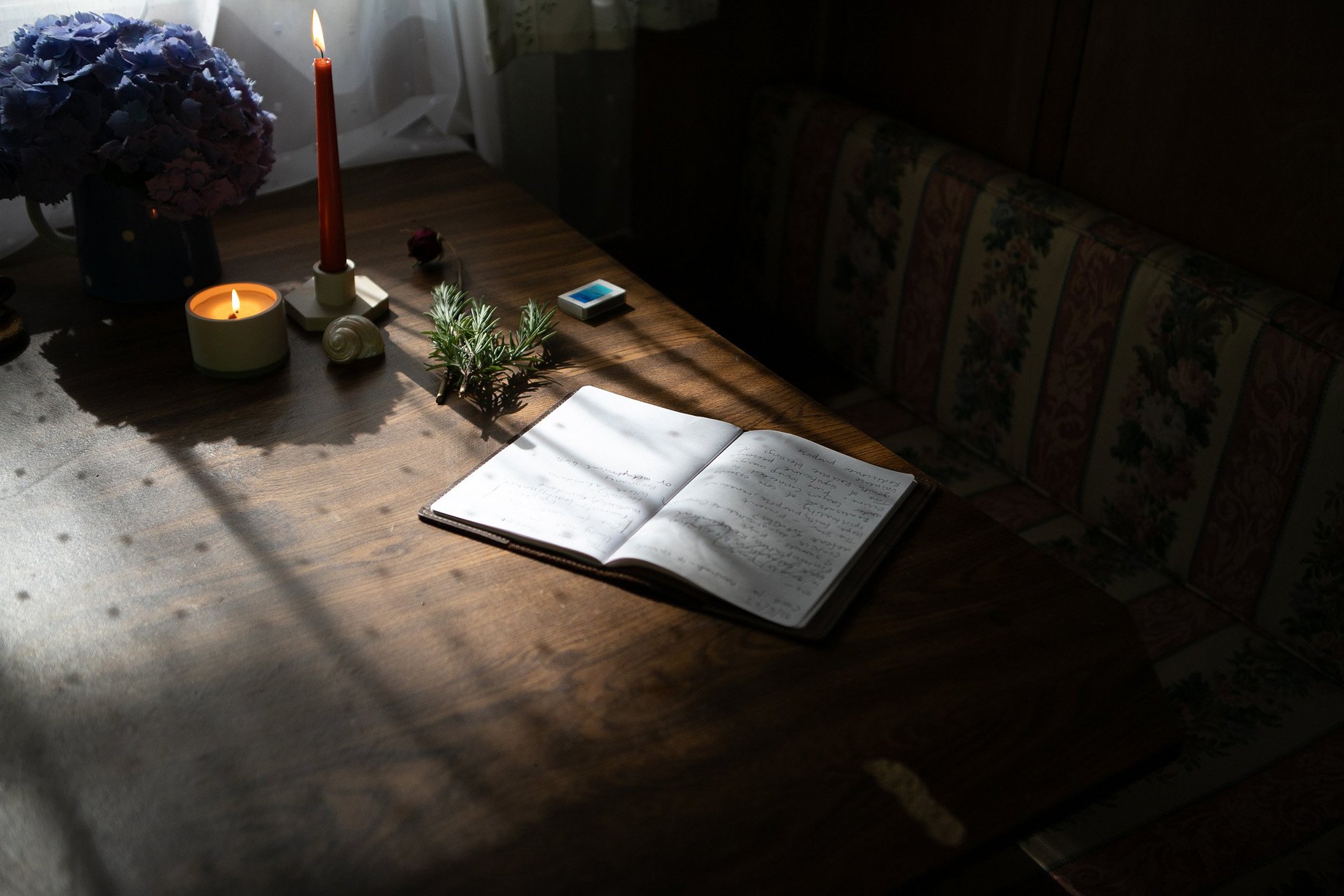 Open journal on a wooden table with candles and herbs in soft window light.