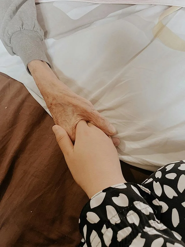 A hand holding an elderly person’s hand on a bed.