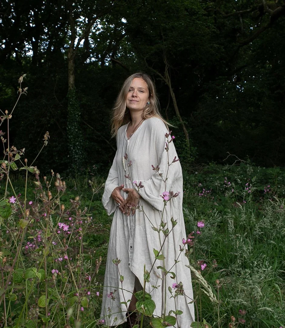Katie Rose in a long, white dress stands in a green, flower-filled outdoor setting with trees in the background.