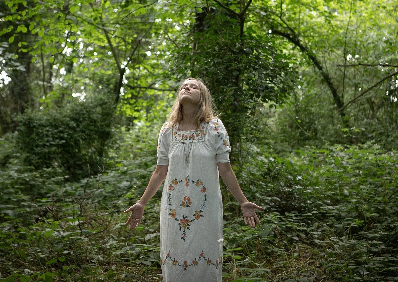 Katie Rose in a white embroidered dress stands in a lush green forest with her arms outstretched and eyes closed, appearing serene and at peace.