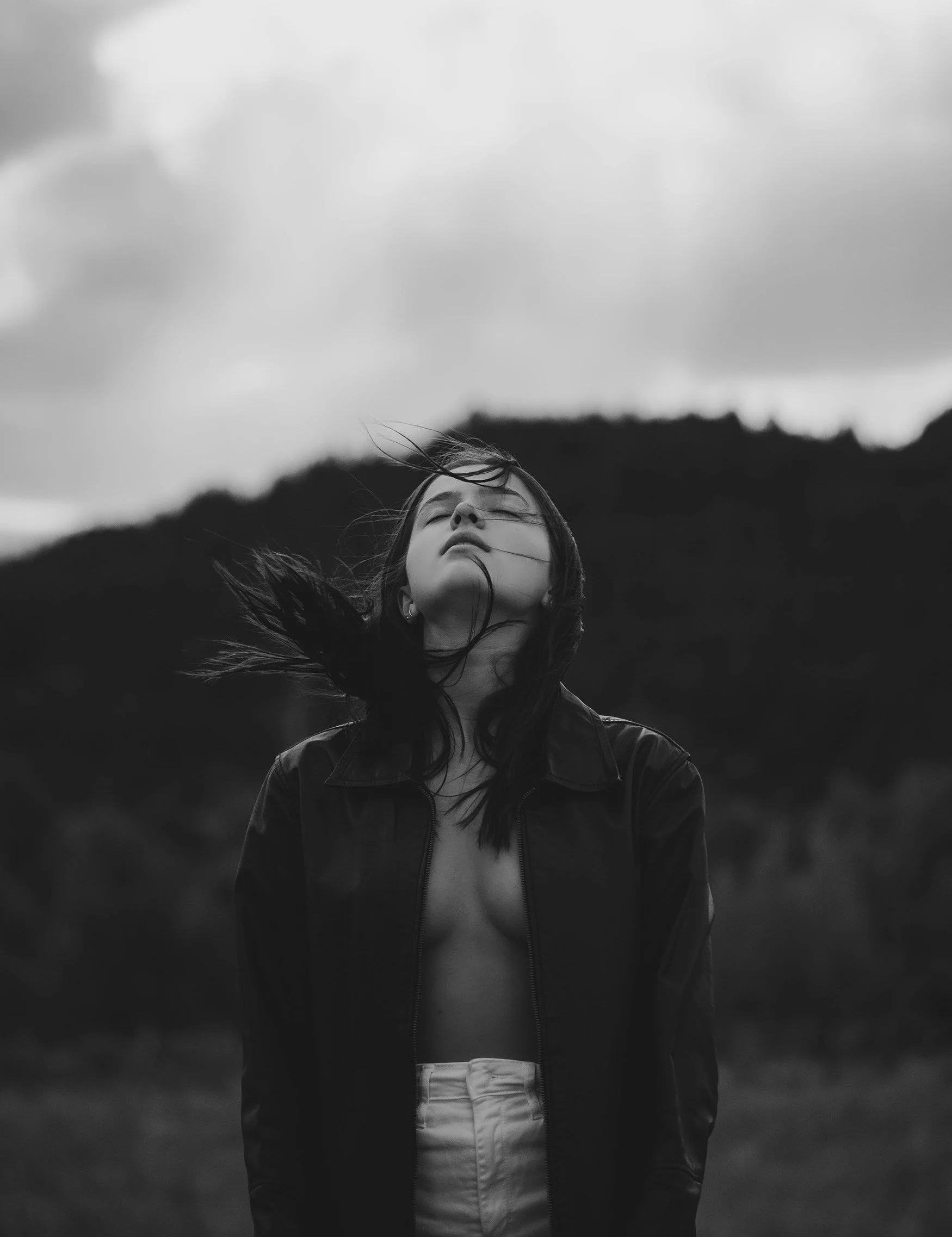 A black and white photo of a woman with long hair, wearing a jacket, standing outdoors with mountains and cloudy sky in the background, with her eyes closed and head tilted back.