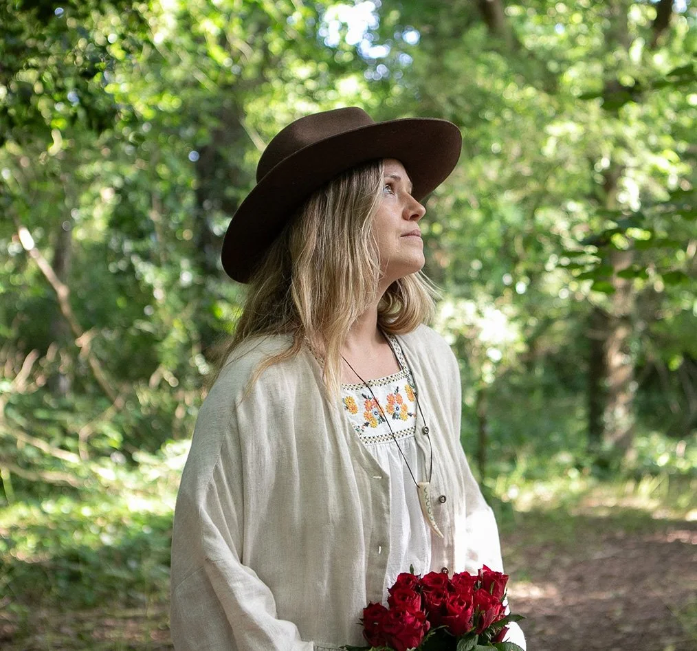 Katie Rose, death doula in Hastings, East Sussex, standing in a woodland setting holding red flowers.