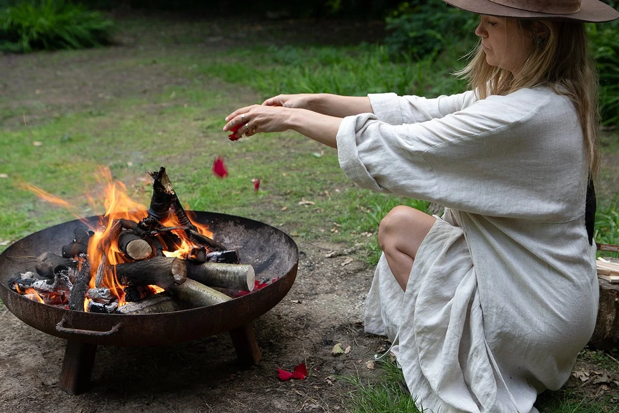Katie Rose sitting outdoors beside a fire bowl, releasing flower petals as part of a grief circle ritual.