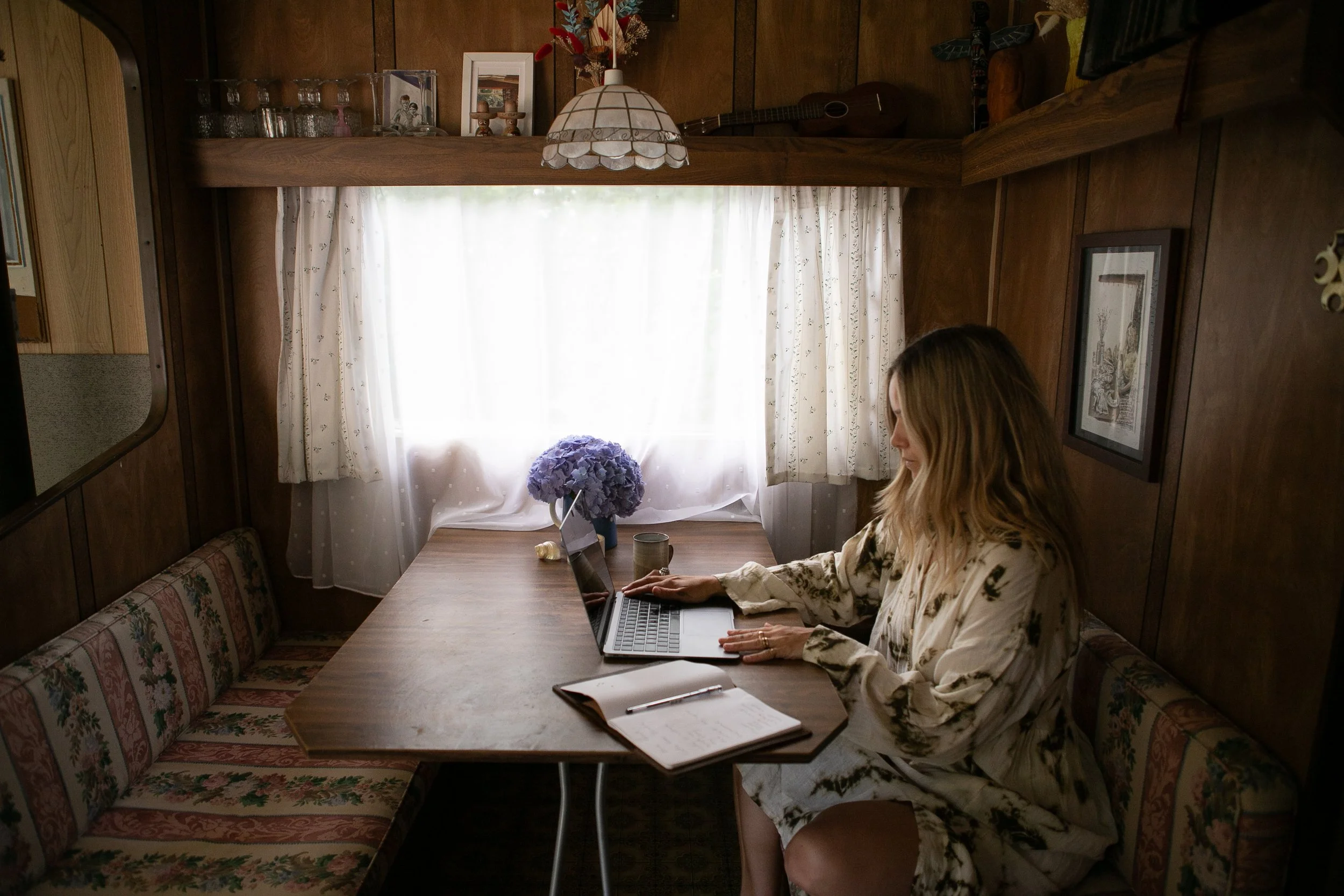 Woman working on a laptop at a wooden table in a cozy, wood-paneled room with a window and floral curtain, decorated with pictures, flowers, and a guitar.