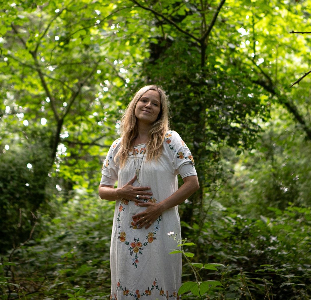 Katie Rose standing in a lush green forest, wearing a white embroidered dress with orange and yellow floral patterns, smiling gently with her hands on her stomach.