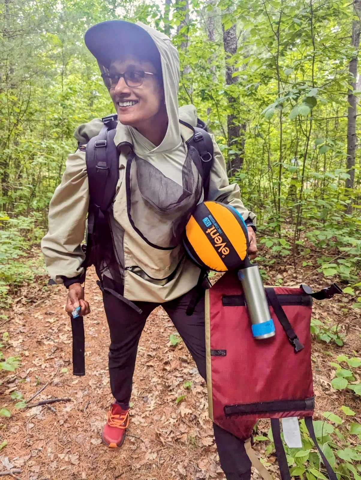 A smiling queer South Indian wearing a backpack and a bug jacket, and carrying a toothbrush, compression sack, travel mug, and folding camp chair.