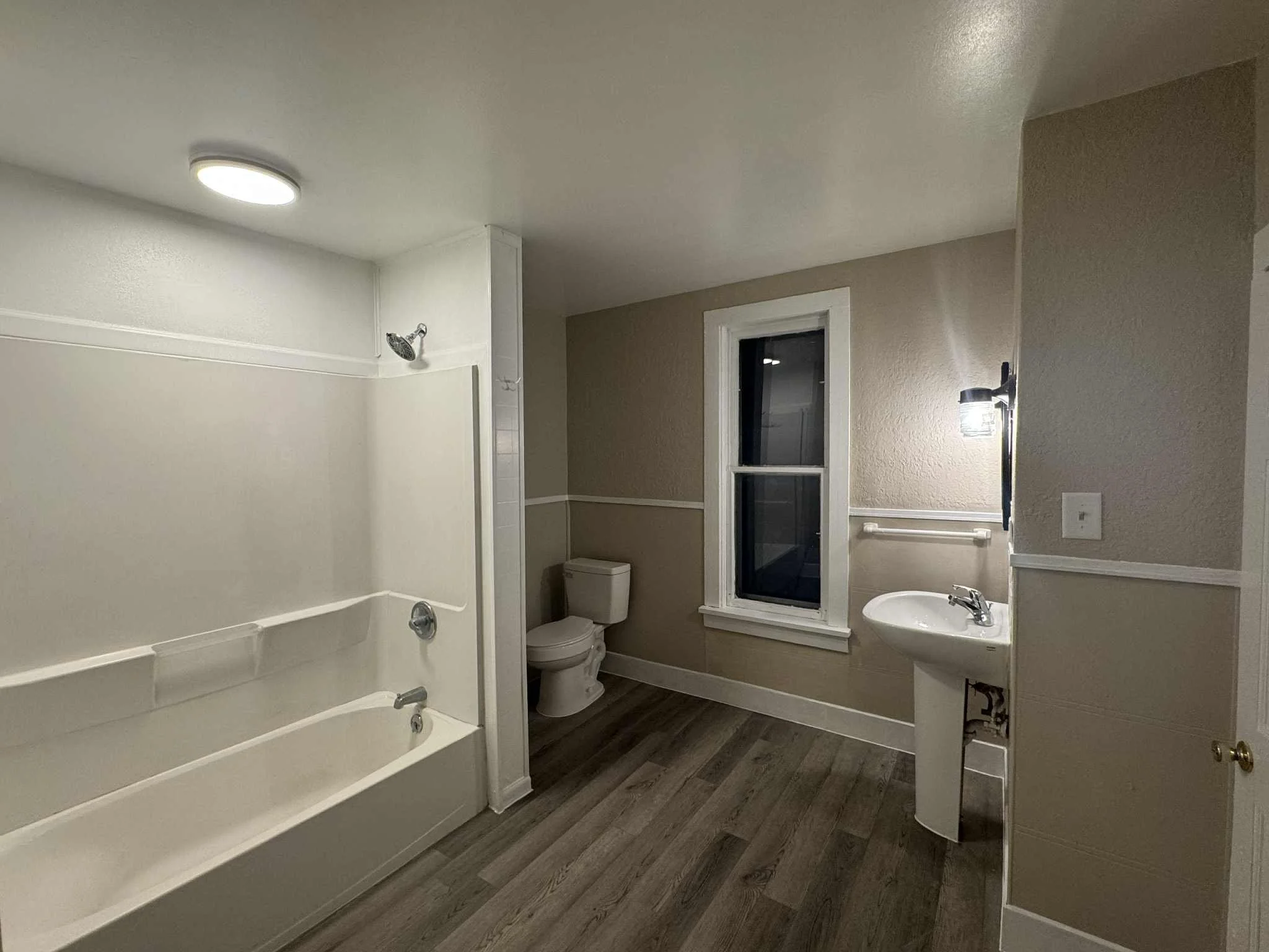 An empty bathroom with a bathtub and shower, toilet, and a small sink near a window with beige walls and wood-look flooring.