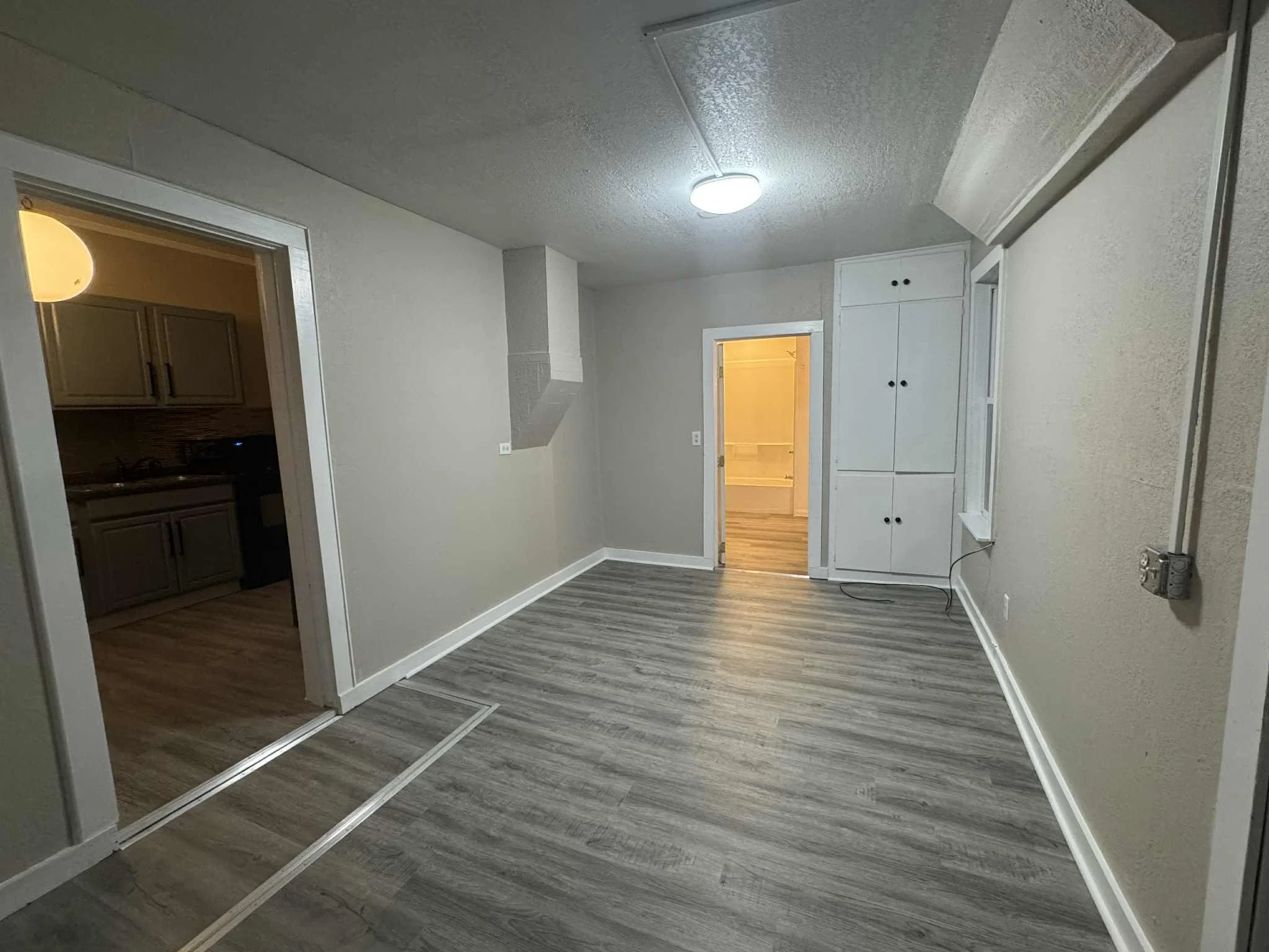 Empty room with wood flooring, white walls, and an open doorway leading to a kitchen with grey cabinets. There is built-in cabinetry on the wall and a light fixture on the ceiling.