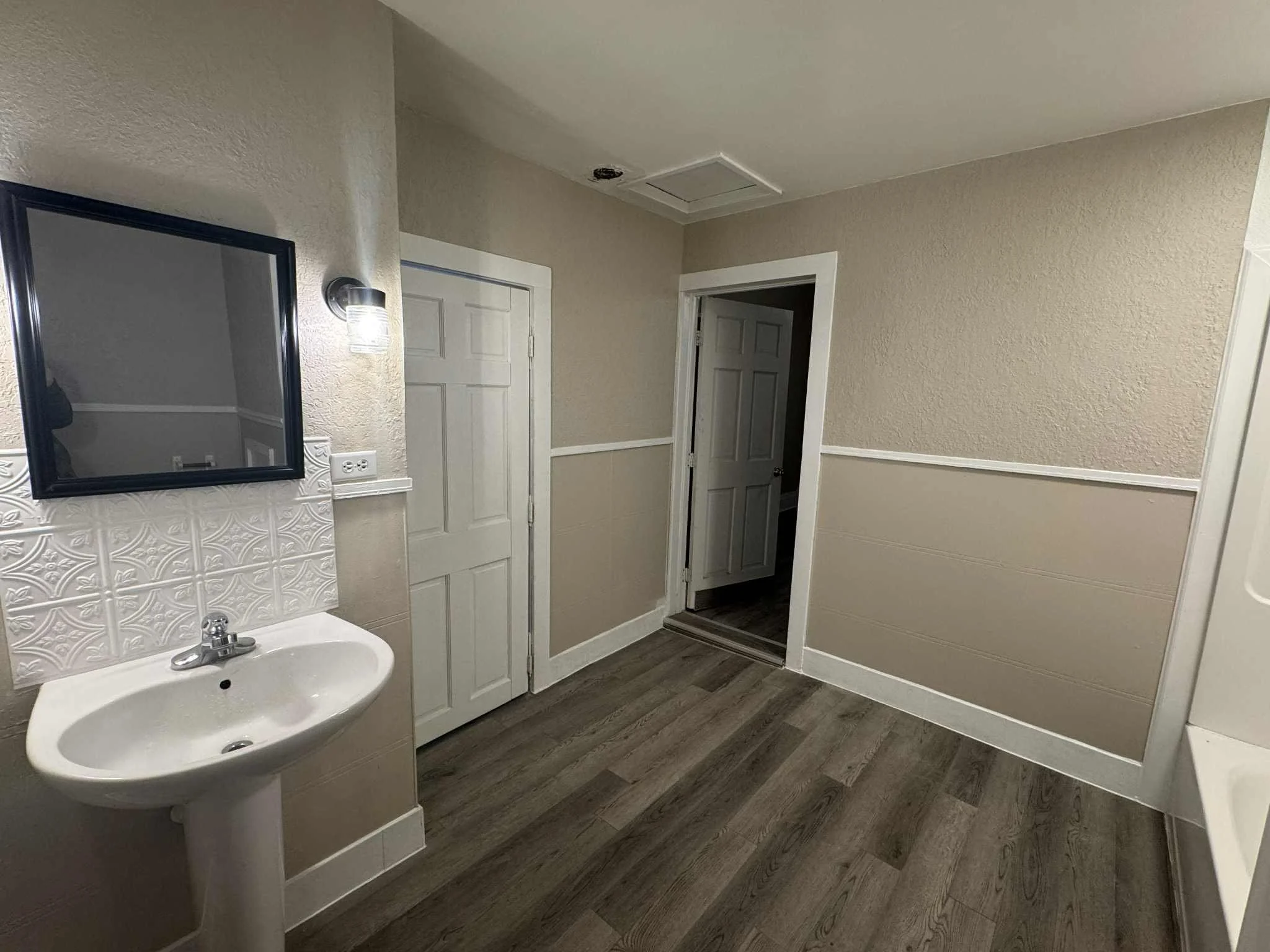 Empty bathroom with small pedestal sink, mirror, wall lamp, beige walls with white trim, wood-look vinyl flooring, and two closed doors.