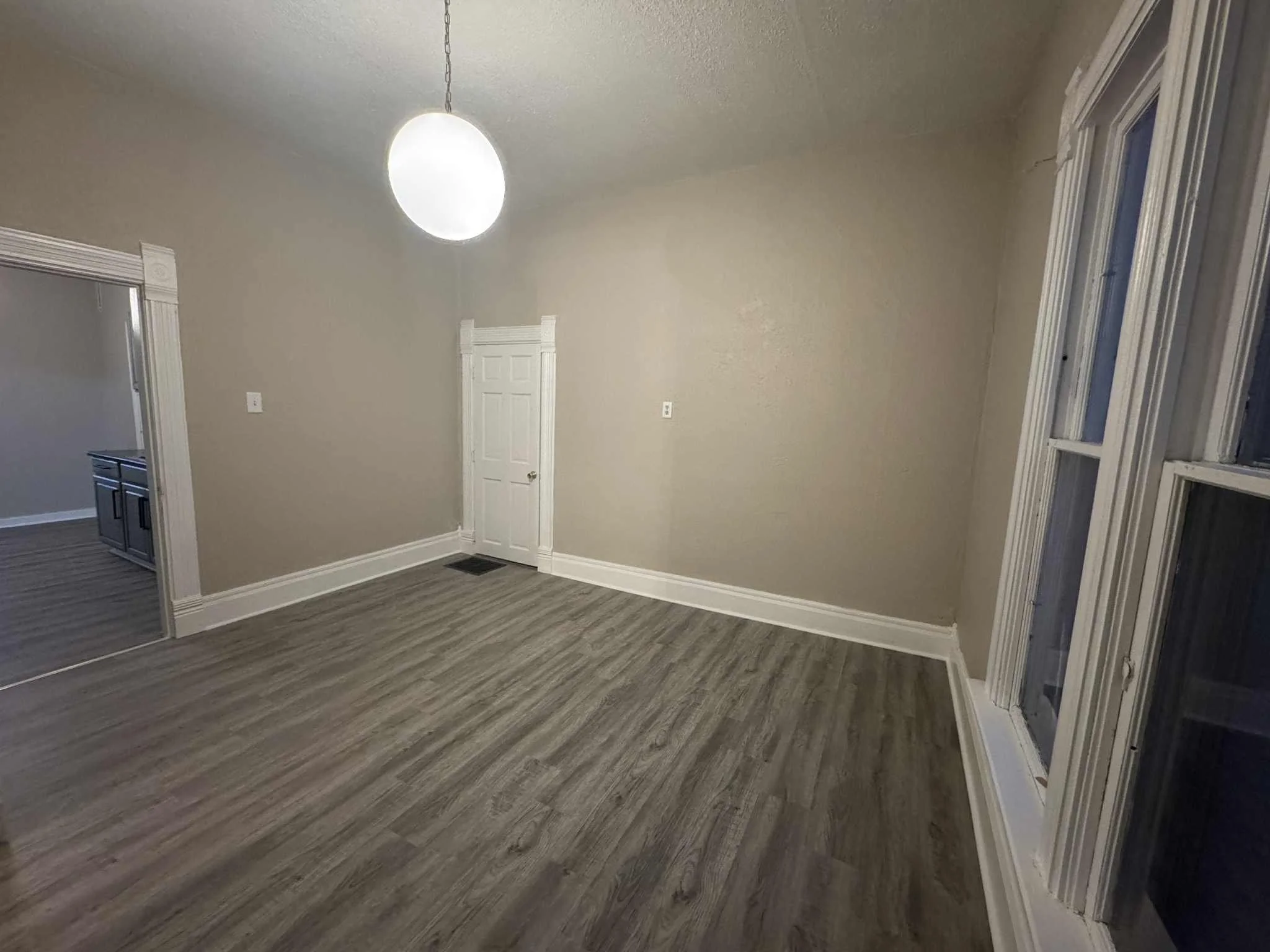 Empty room with beige walls, white trim, a ceiling light, and windows on the right, with a doorway leading to another room on the left.