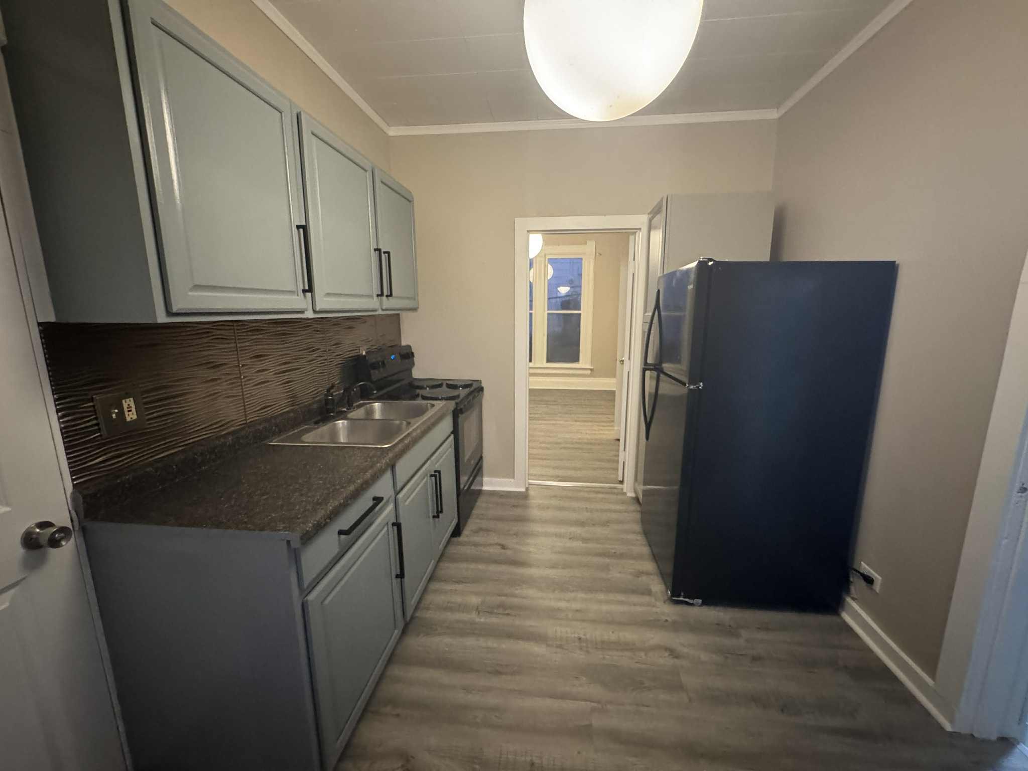 Kitchen with gray cabinets, black countertops, a stainless steel sink, a stove, and a black refrigerator. Light-colored walls and wood-look flooring.