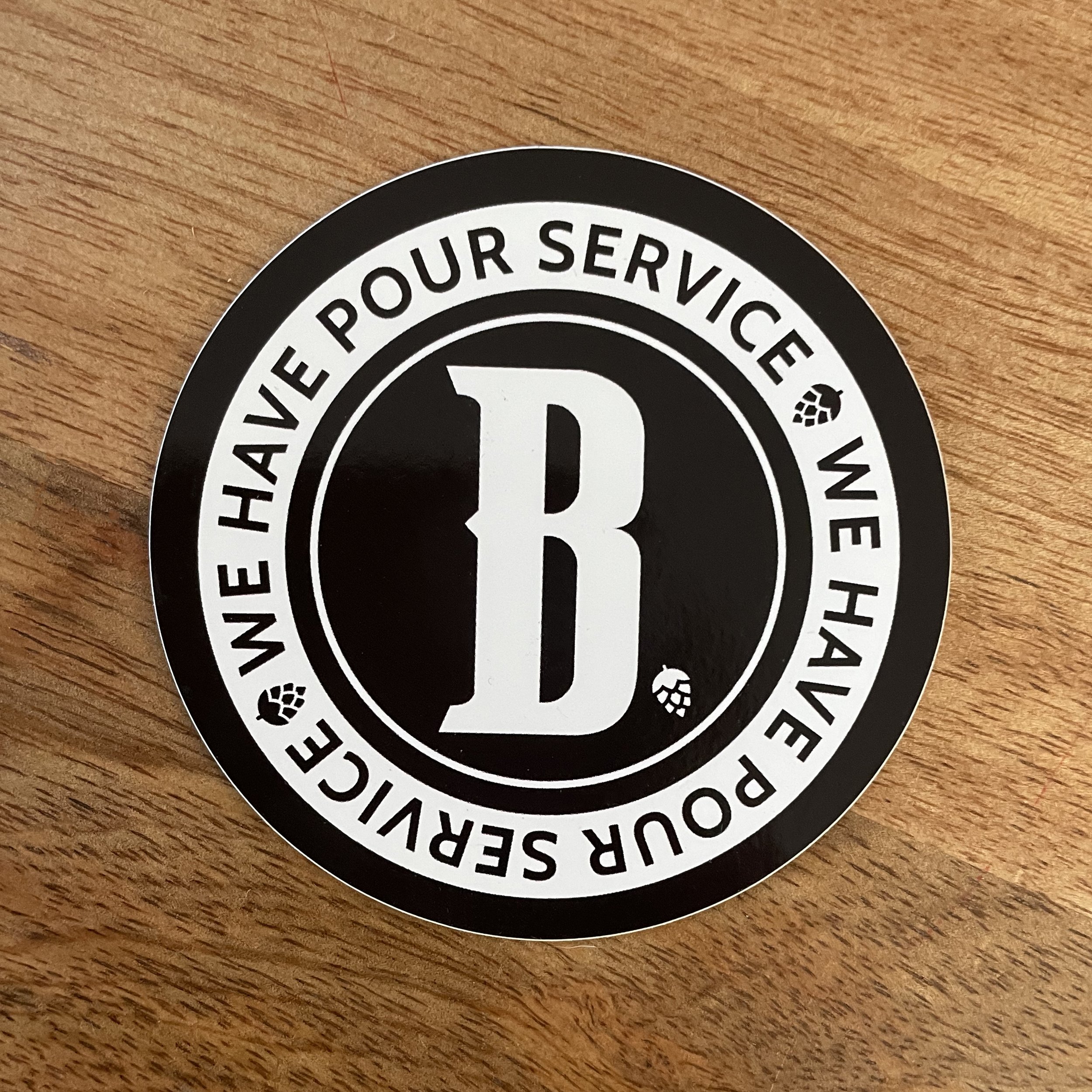 Black and white circular sticker with large letter 'B' in the center, surrounded by the text 'WE HAVE POUR SERVICE' repeated twice with small hop icons between the words, on a wooden surface.
