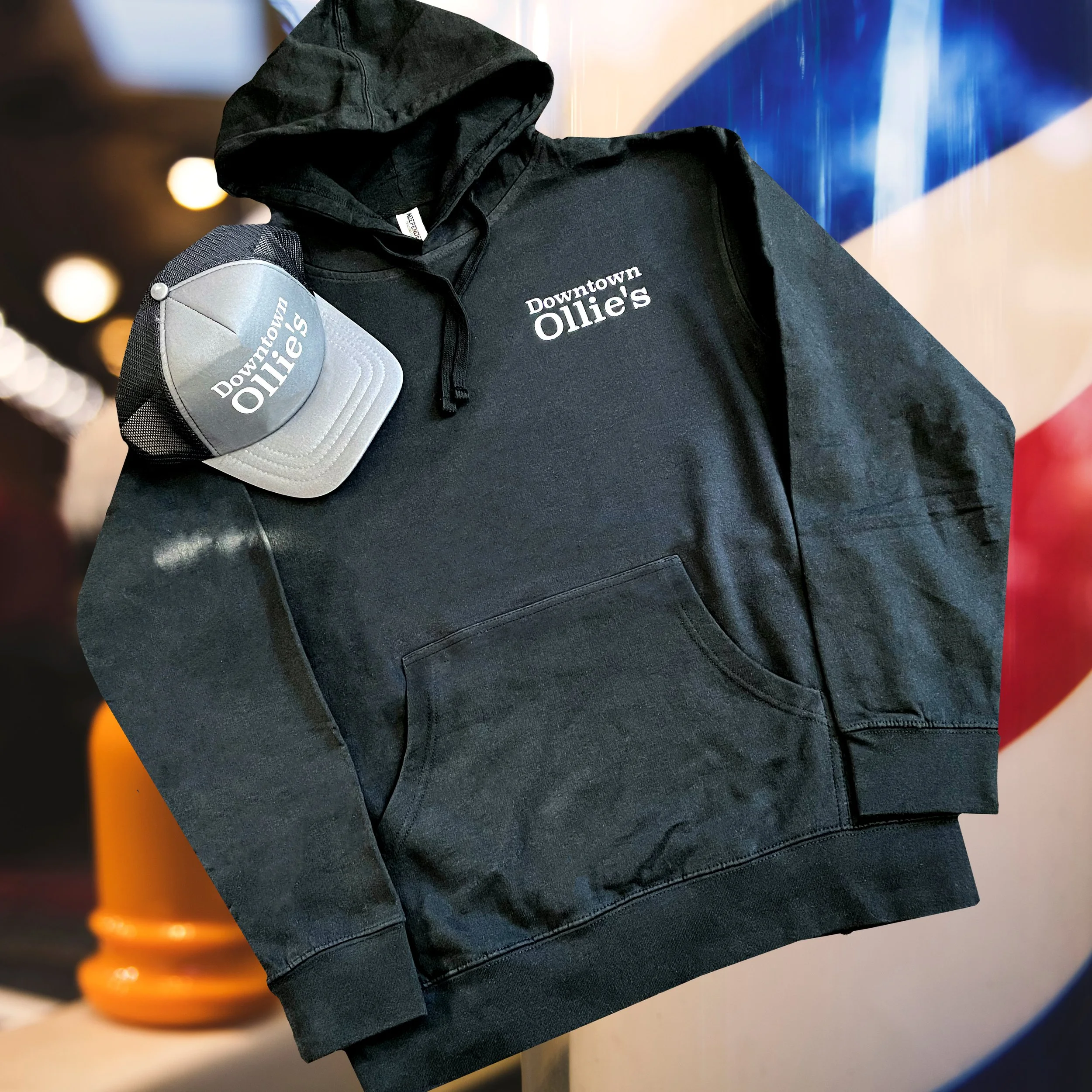 Black hoodie with white embroidered text 'Downtown Ollie's' and a matching cap with the same text, set against a blurred background.