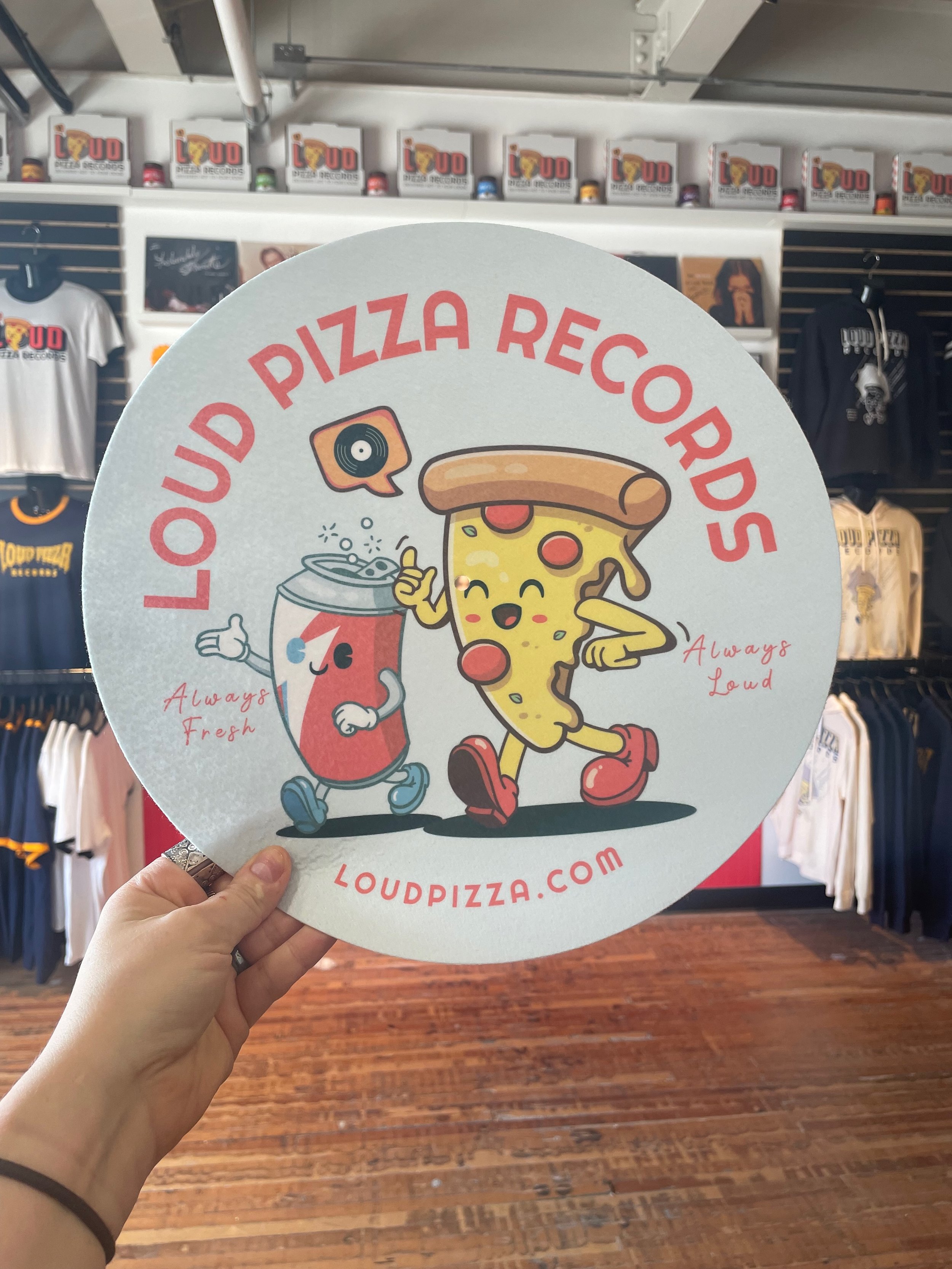 A circular sign with cartoon characters of pizza and a soda cup, saying "Loud Pizza Records" with the website loudpizza.com, in a store with T-shirts and memorabilia in the background.