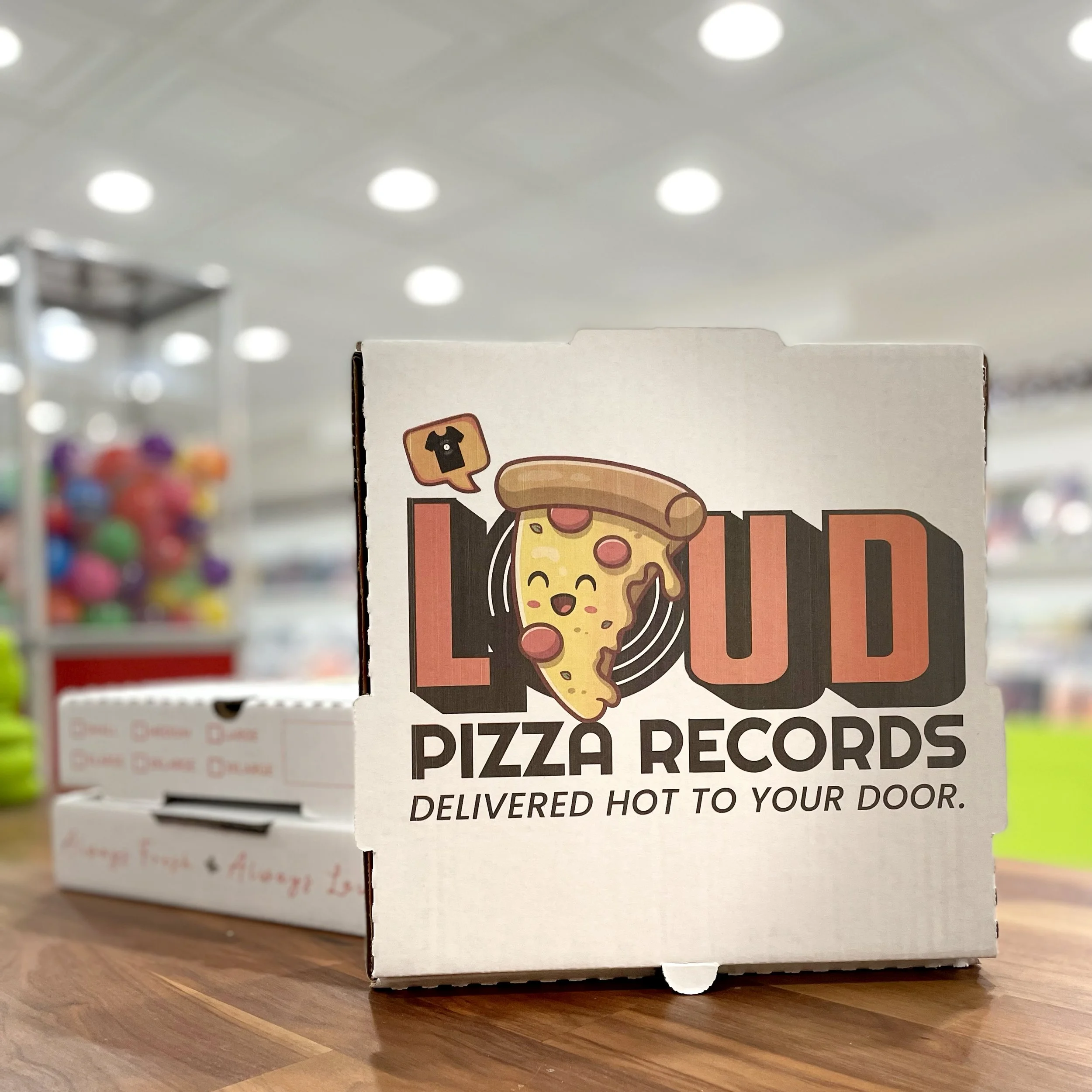 Pizza box with a cartoon slice of pizza featuring a happy face and a pizza tower. Text on the box reads 'LOUD PIZZA RECORDS' with a slogan 'Delivered hot to your door.' in a store setting.